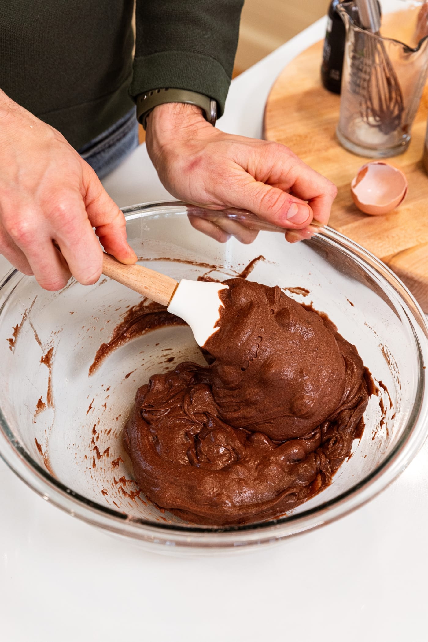 Hand mixing chocolate batter for chocolate cake with white spatula in glass bowl on white surface.