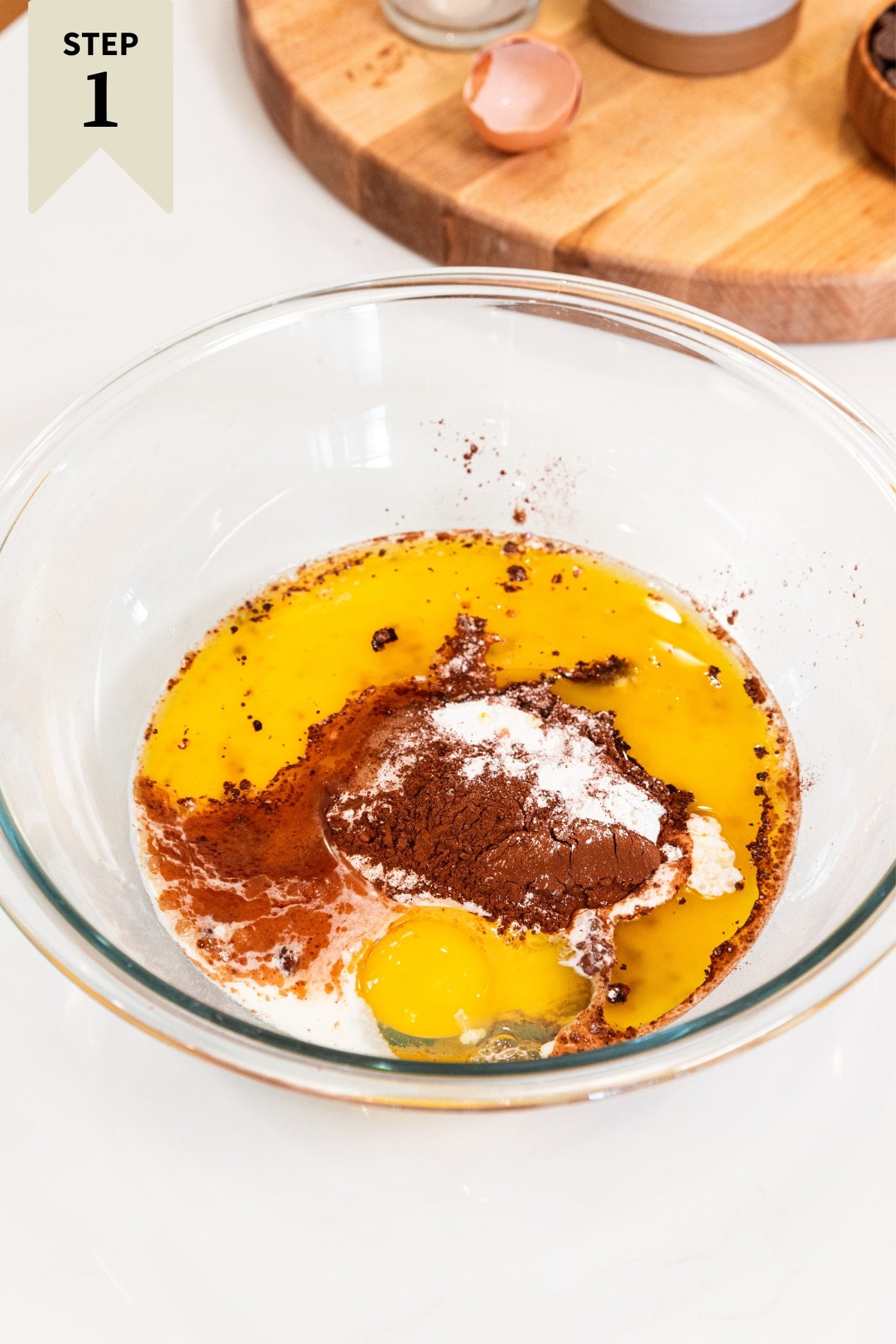 Glass bowl sitting on white countertop filled with all ingredients needed for chocolate cake including cocoa powder, sugar, egg, milk, and more.