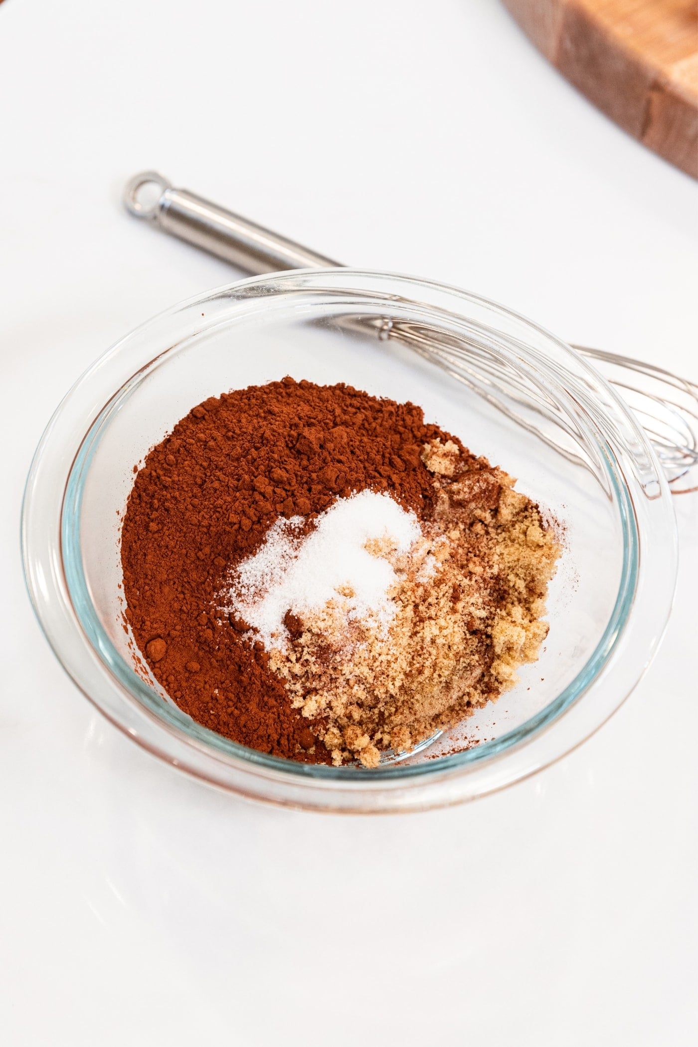 Glass bowl filled with cocoa powder, sugar, and brown sugar sitting on white countertop before being mixed with whisk.