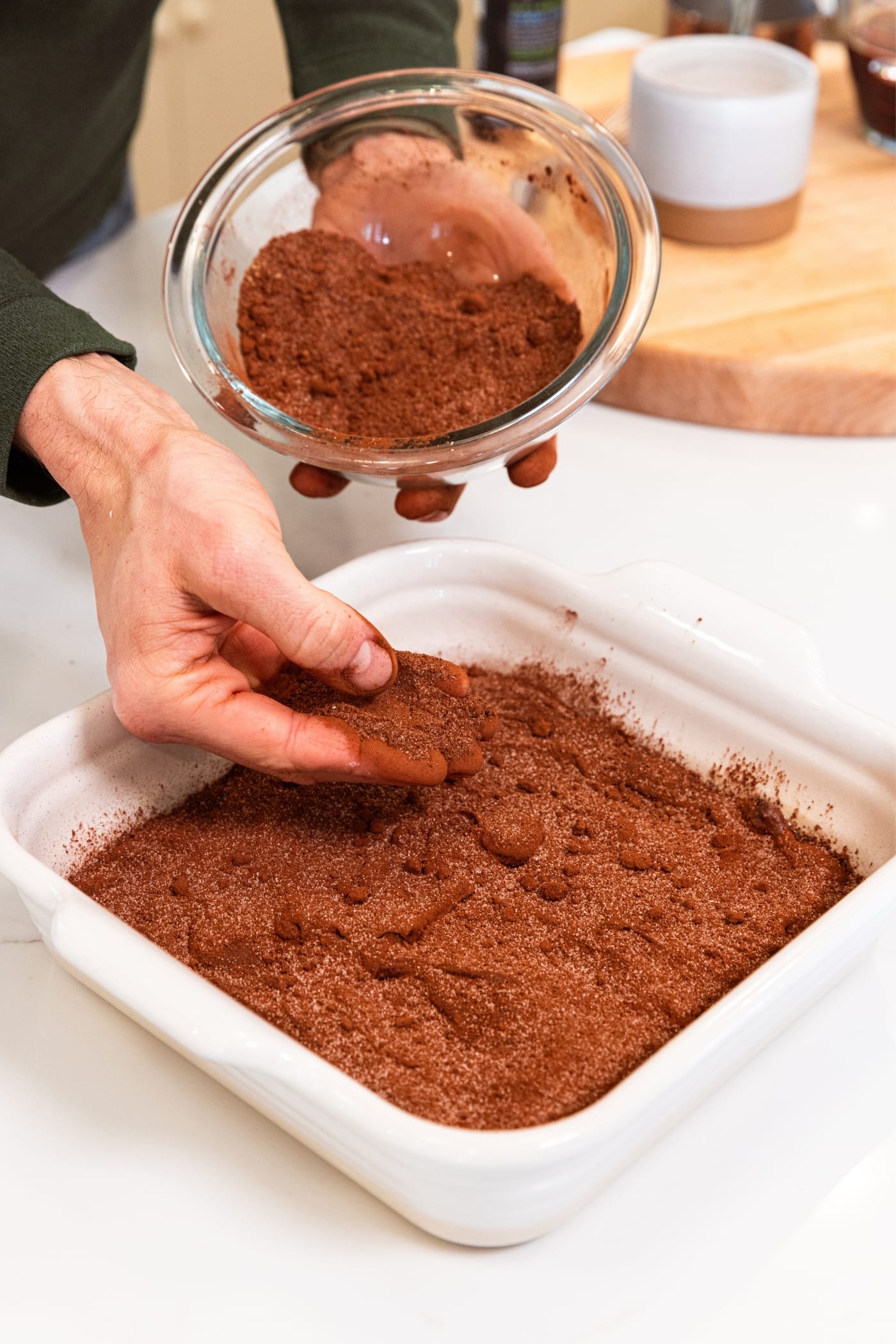 Hand sprinkling chocolate sugar mixture on top of chocolate batter in white baking dish sitting on white countertop.