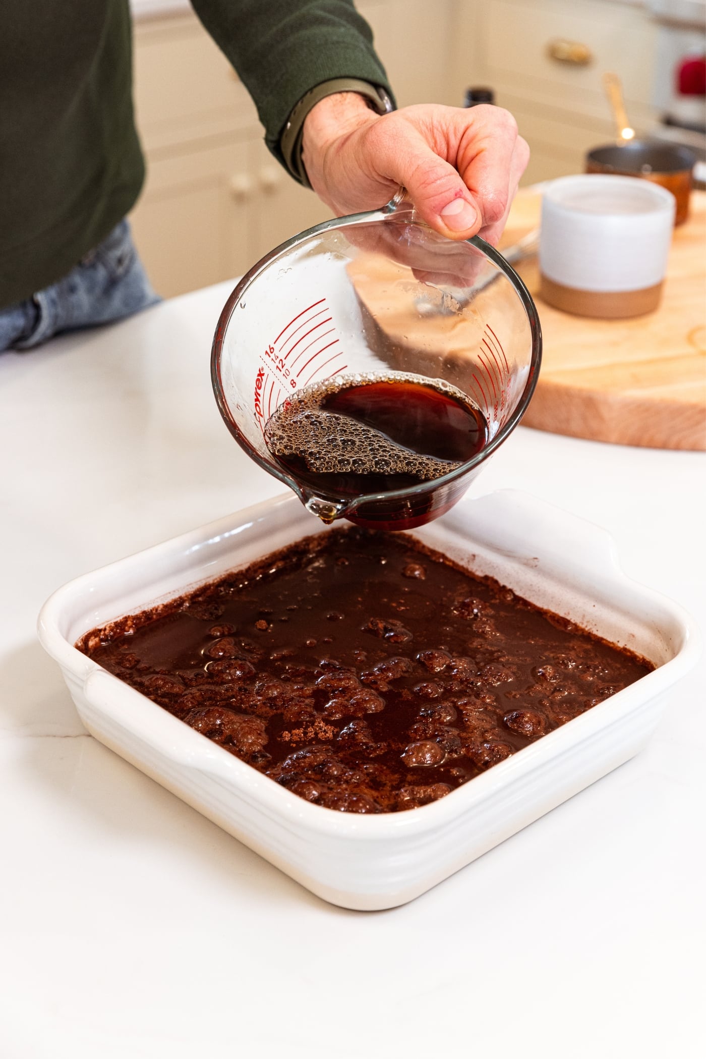Hand using glass measuring glass to pour coffee on top of chocolate batter topped with brown sugar and cocoa mixture.