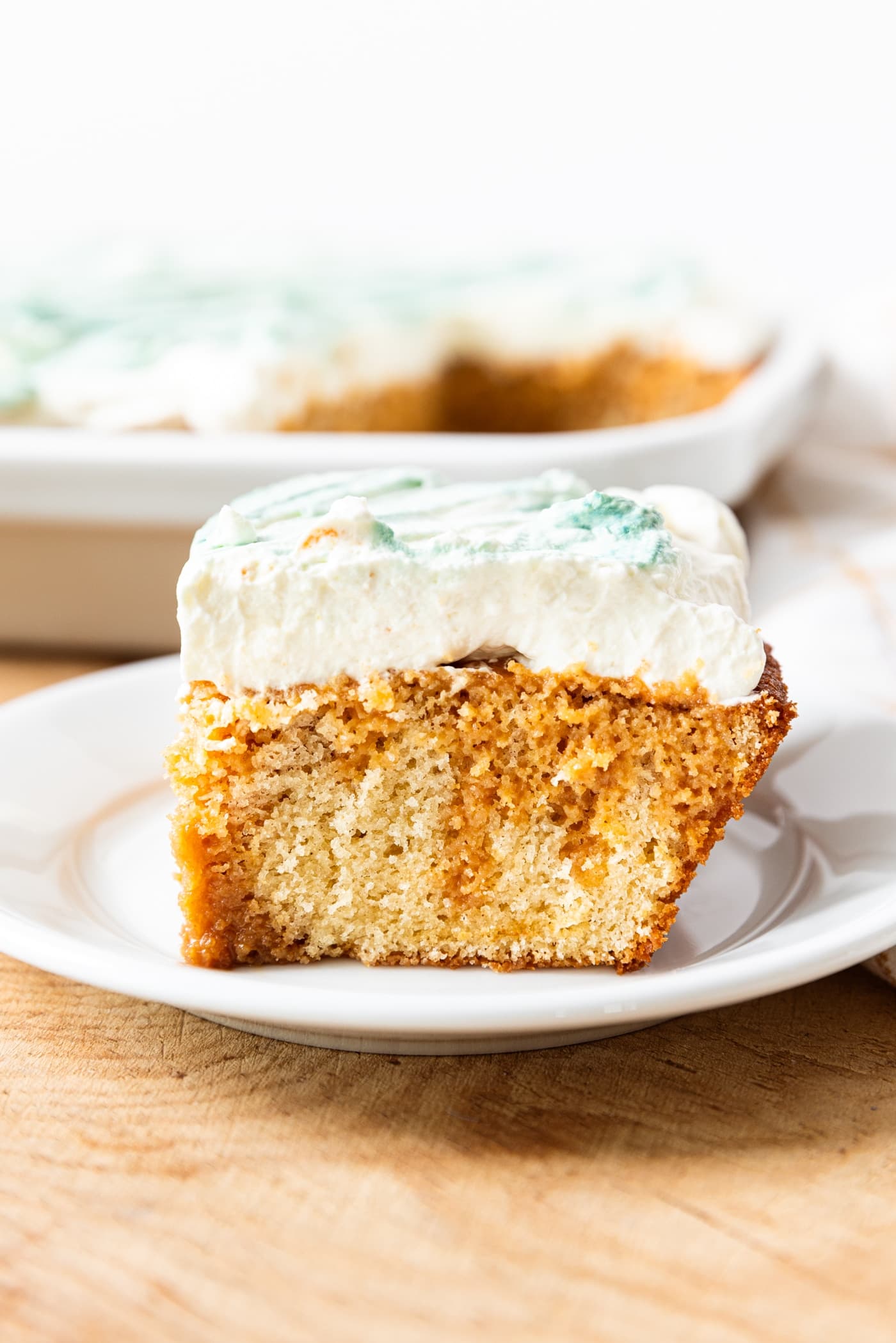 Piece of tres leches cake showing two different colors of cake with whipped topping sitting on white plate on wood surface with rest of cake in the background.