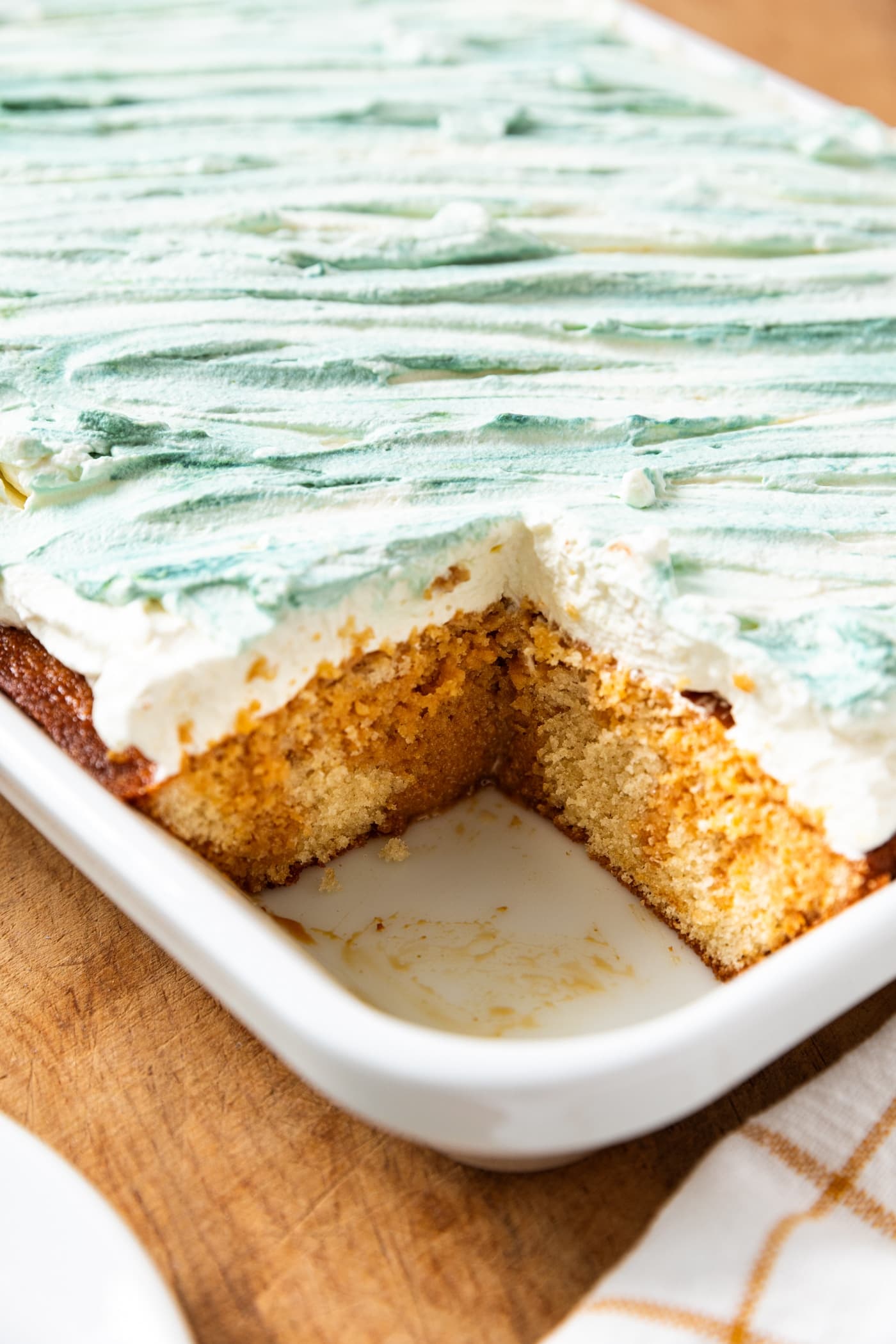 Large baking dish filled with baked tres leches cake with one pieces removed showing the interior texture and colors of the cake with whipped topping spread all over the top.