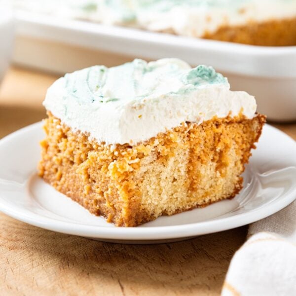 White plate with piece of tres leches cake topped with whipped cream topping with rest of cake in the background all on wooden surface.