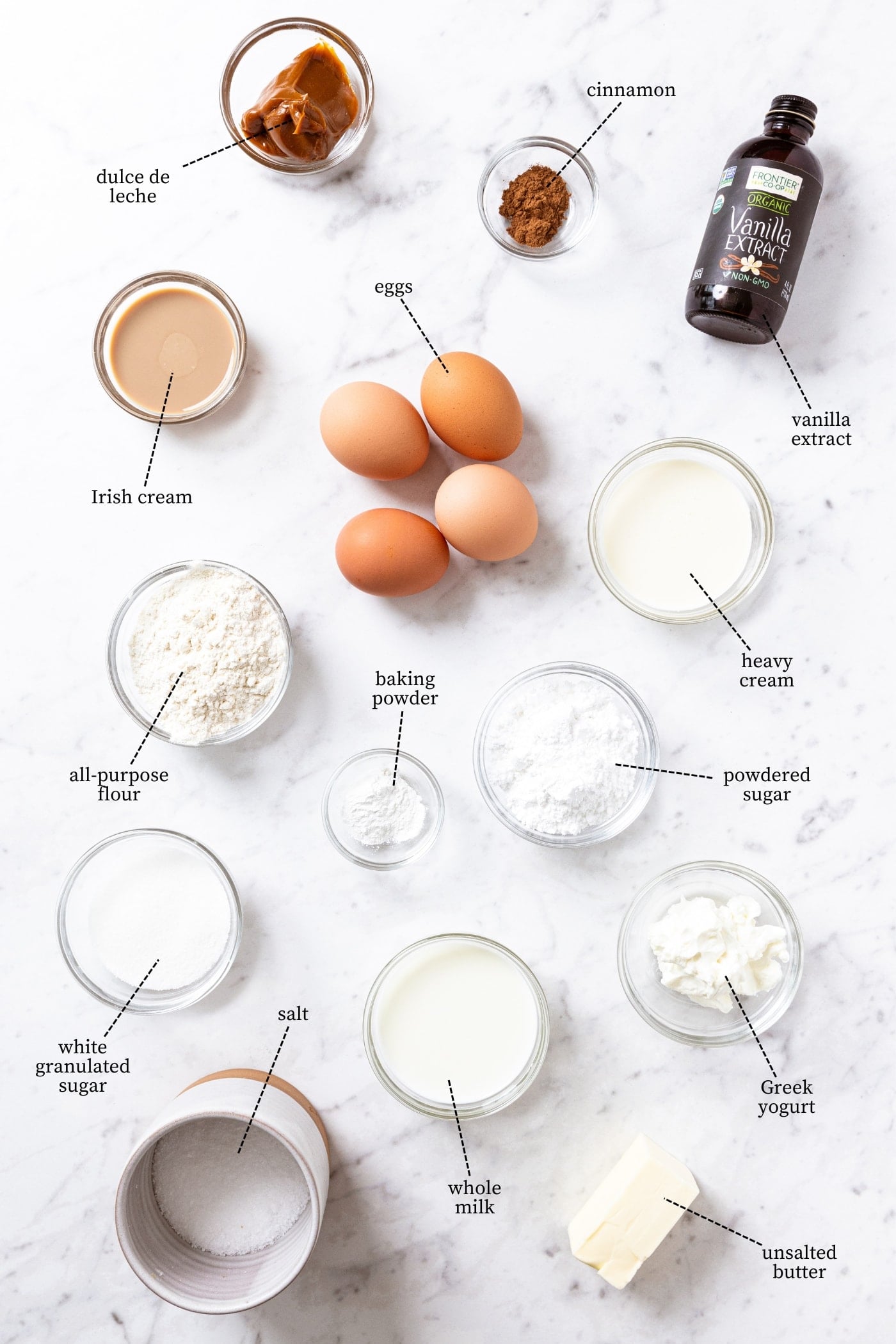 White marble surface filled with all ingredients needed to make tres leches cake including eggs, flour, sugar, vanilla extract, cinnamon, milk, cream, and more.