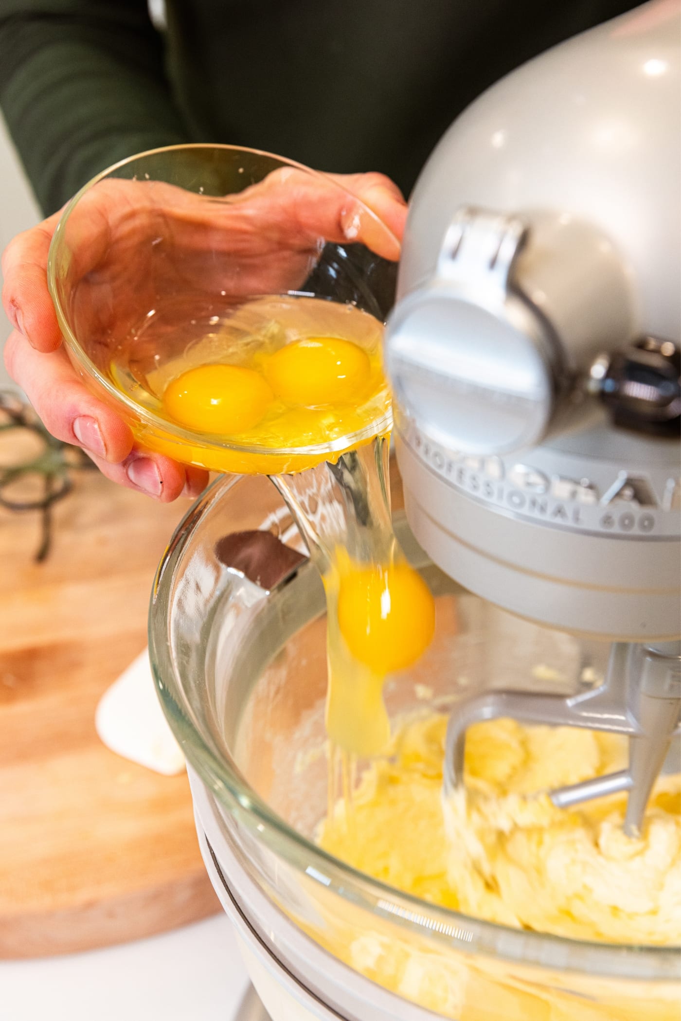 Hand holding small glass bowl filled with cracked eggs pouring one egg at a time into a large glass bowl on stand mixer with batter.