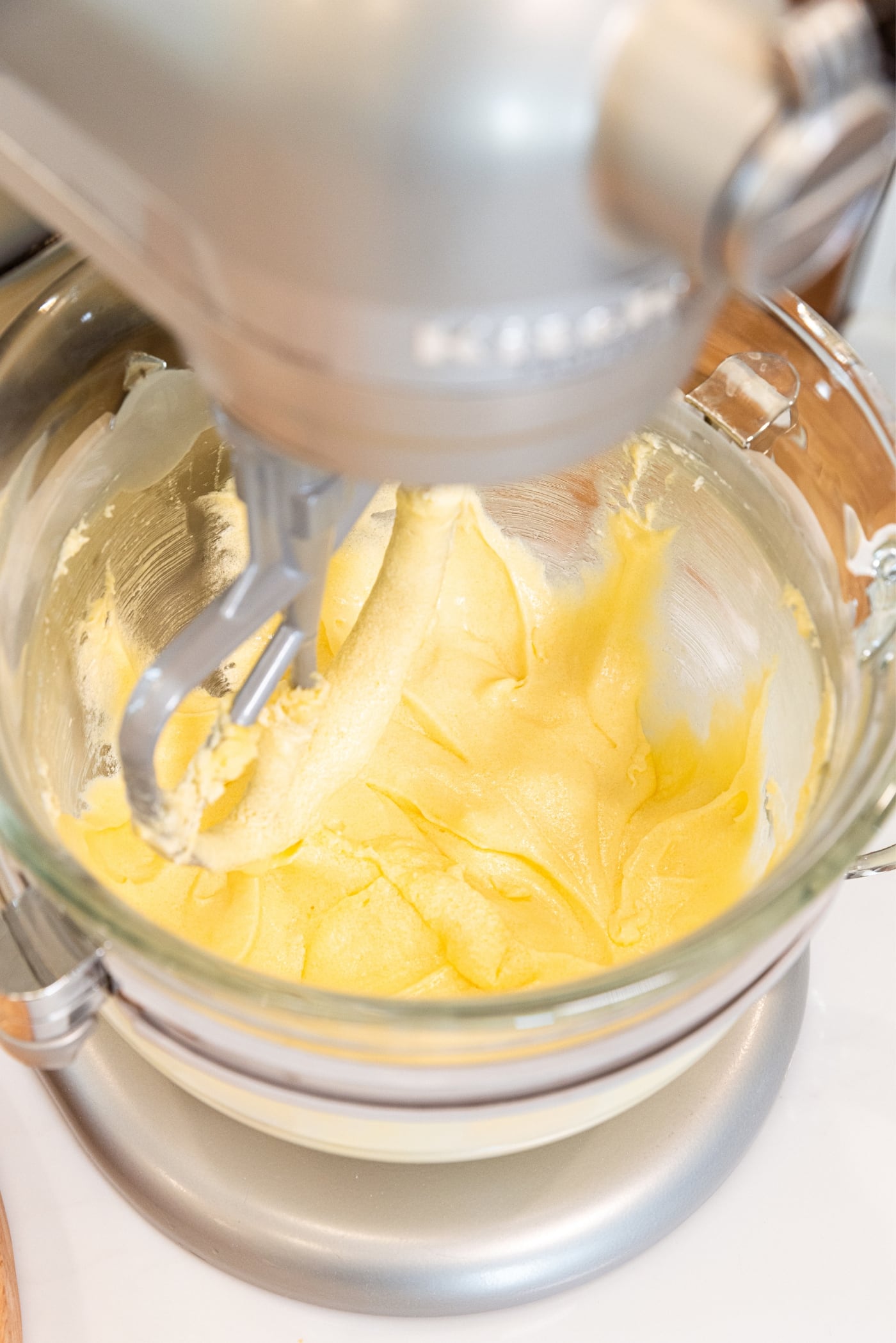 Glass bowl on stand mixer mixing together smooth liquid made of sugar, butter, and eggs all on white countertop.