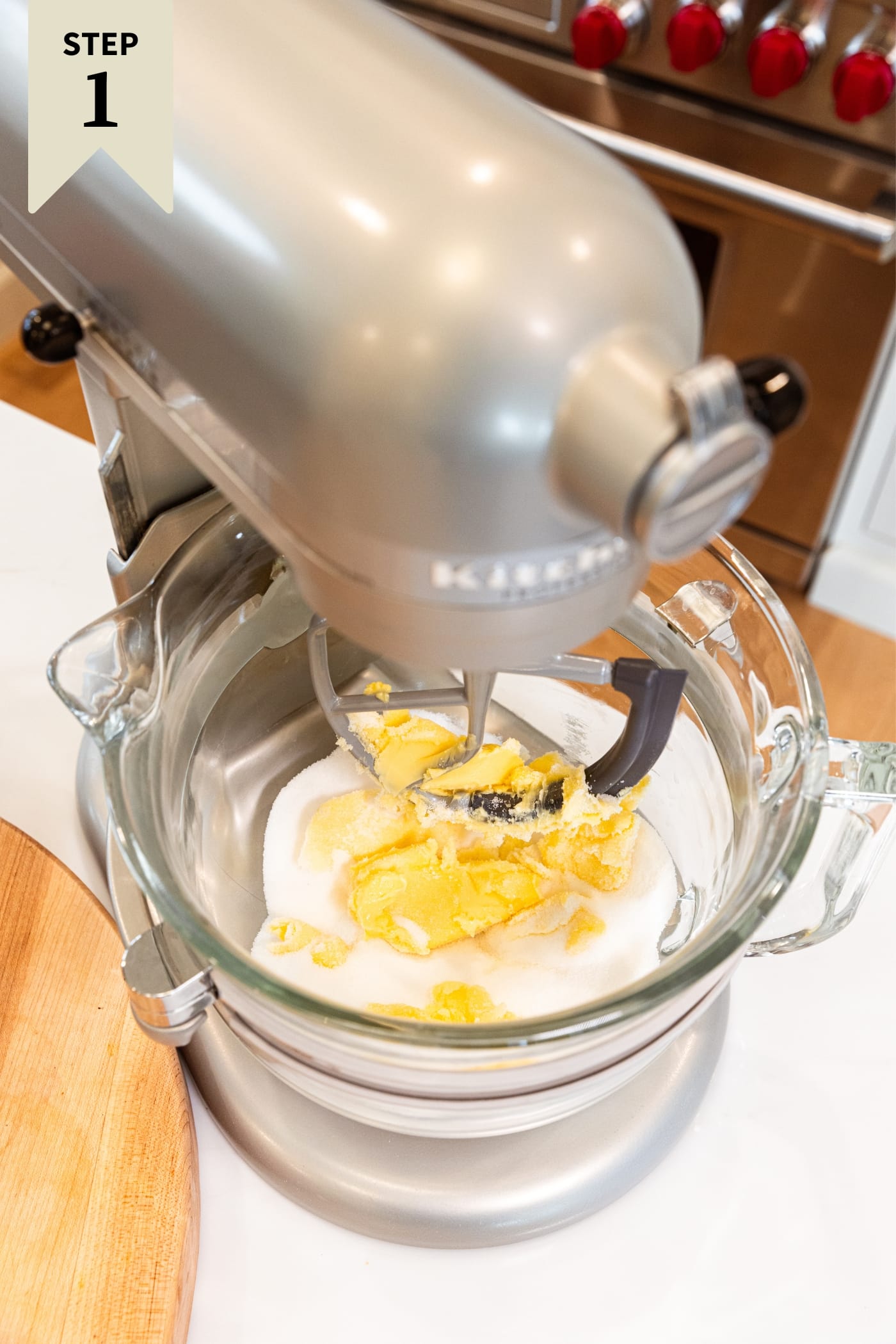 Glass bowl attached to stand mixer mixing together butter and sugar all on white countertop beside wood cutting board.