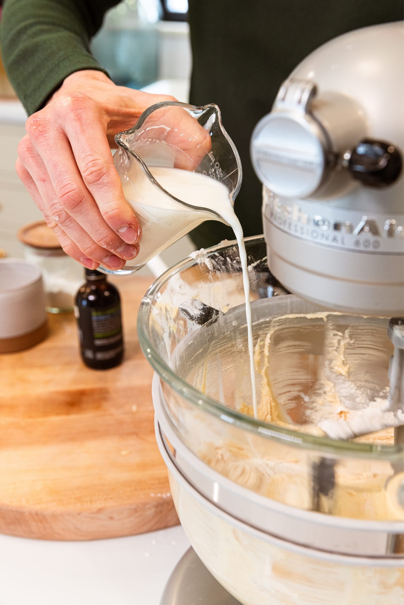 Hand holding glass measuring cup filled with milk pouring some milk into the glass bowl on a stand mixer with flour and batter already in bowl.
