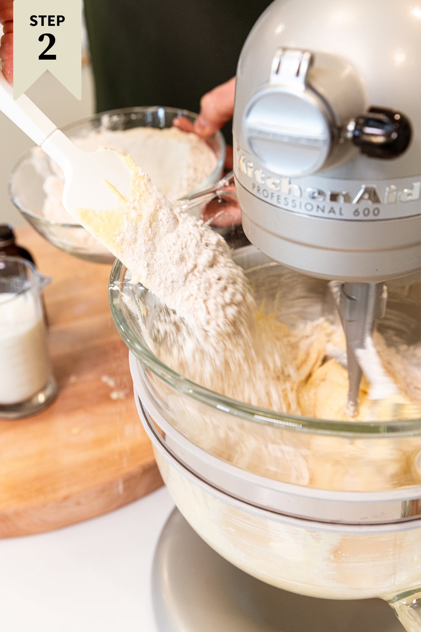 Hand holding small glass bowl filled with flour mixture pushing some of the flour into a large glass mixing bowl on stand mixer with batter already in the bowl.
