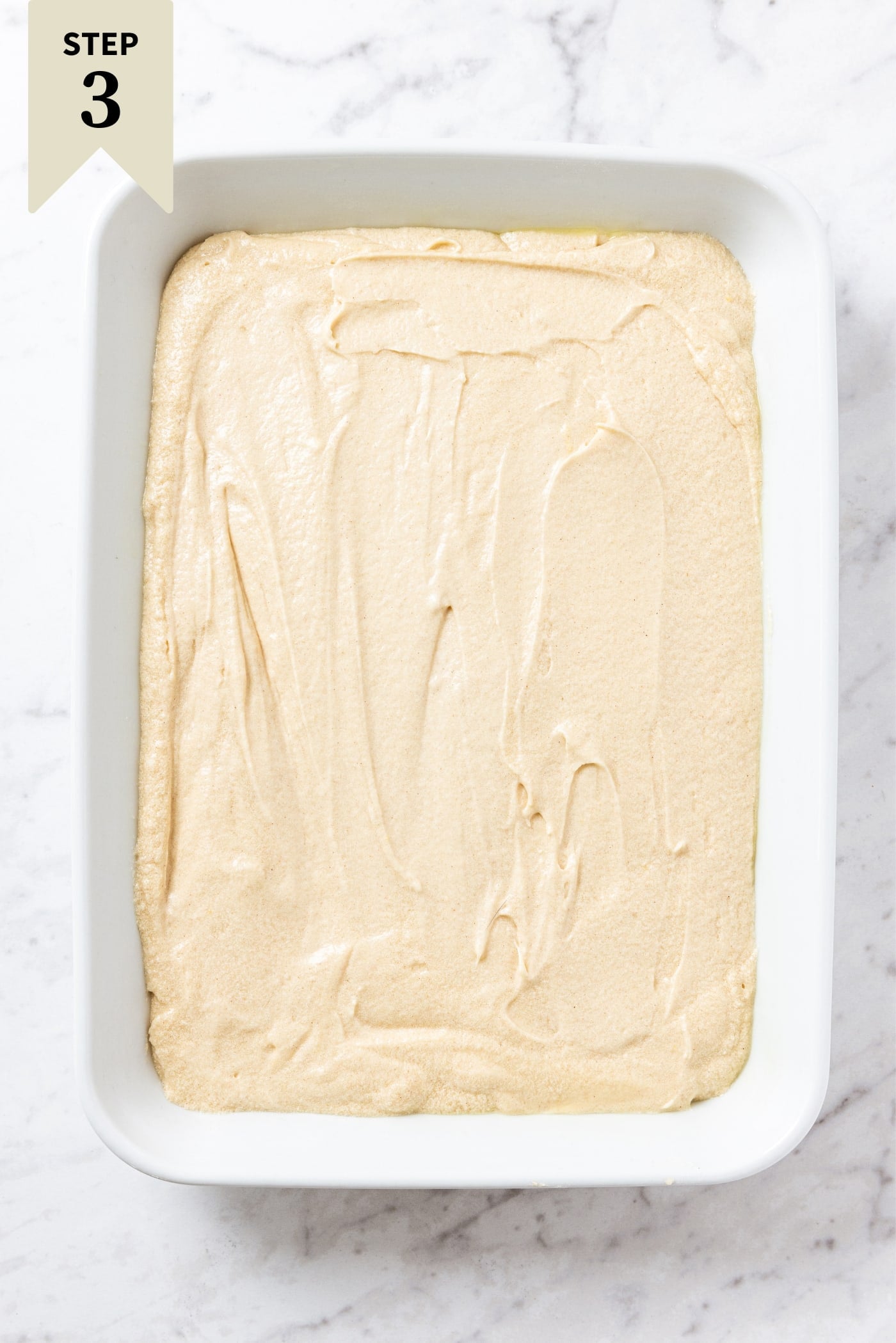 Baking dish with unbaked batter spread out into the white pan all on white marble surface.