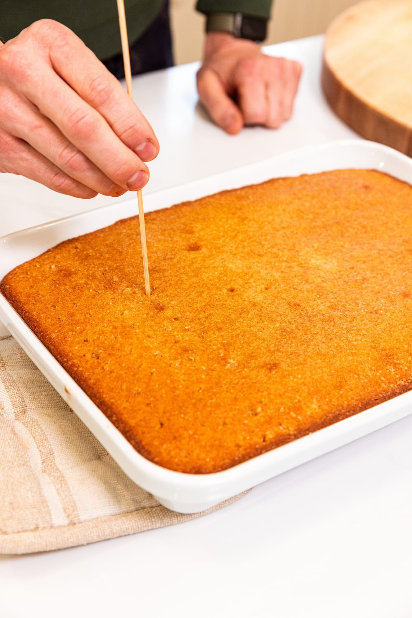 Hand using skewer to poke holes into a large tres leches cake in a white baking dish with golden top on white surface.