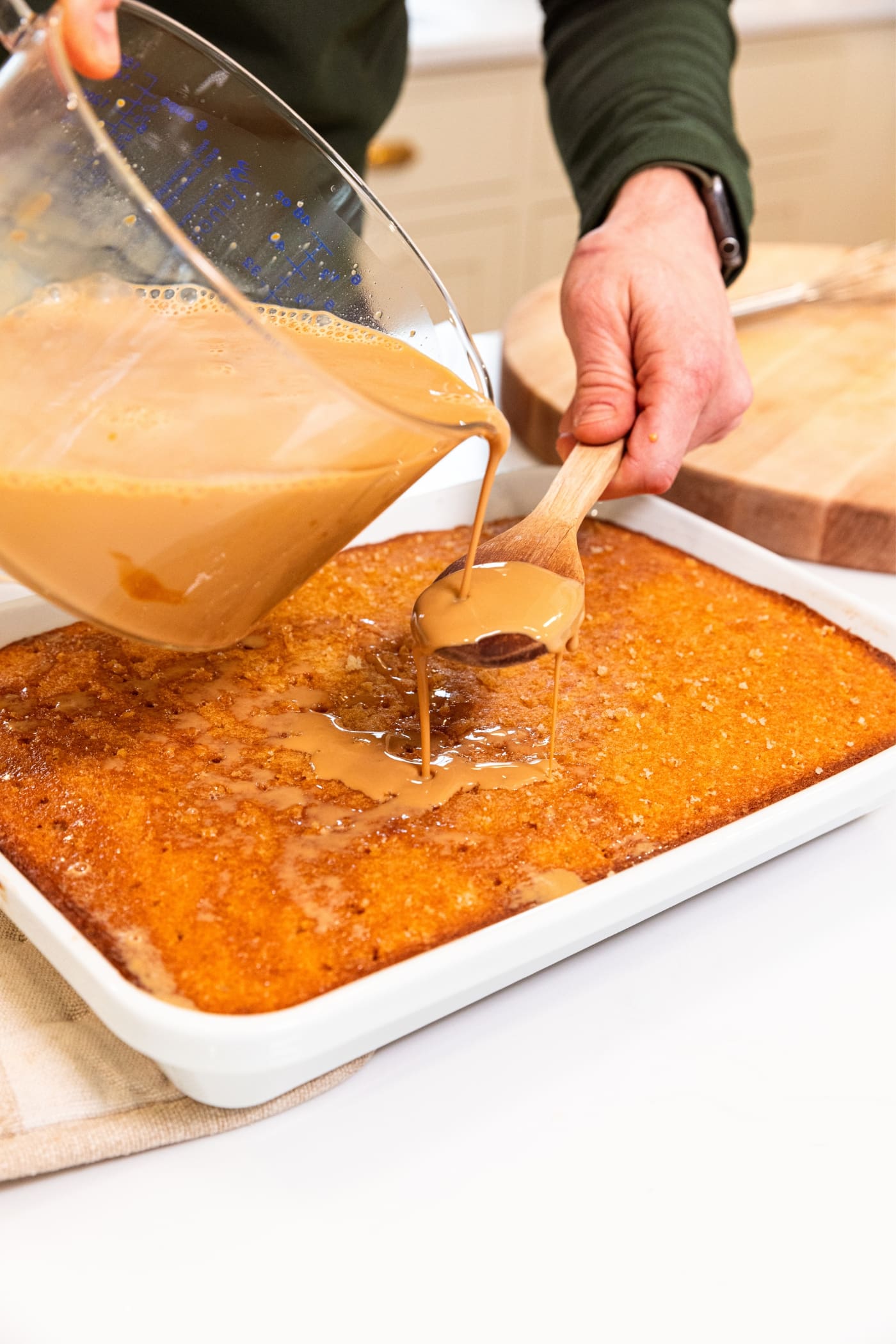 Hand holding wooden spoon pouring dulce de leche cream mixture from large glass measuring cup over the spoon onto the top of a baked tres leches cake in baking dish.