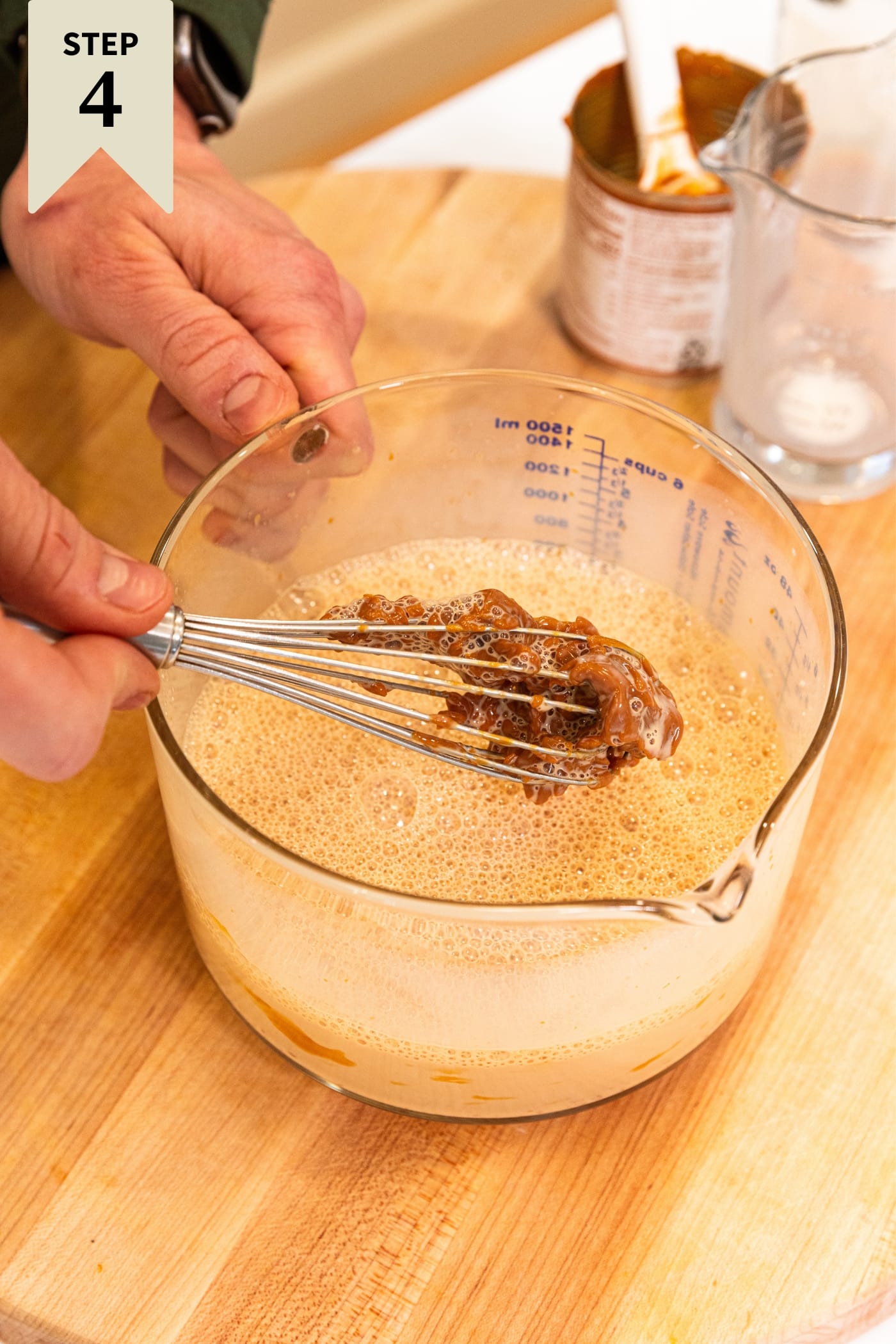 Large glass measuring cup filled with brown colored liquid with hand holding up whisk showing the dulce de leche in the mixture before being mixed fully on wood cutting board.