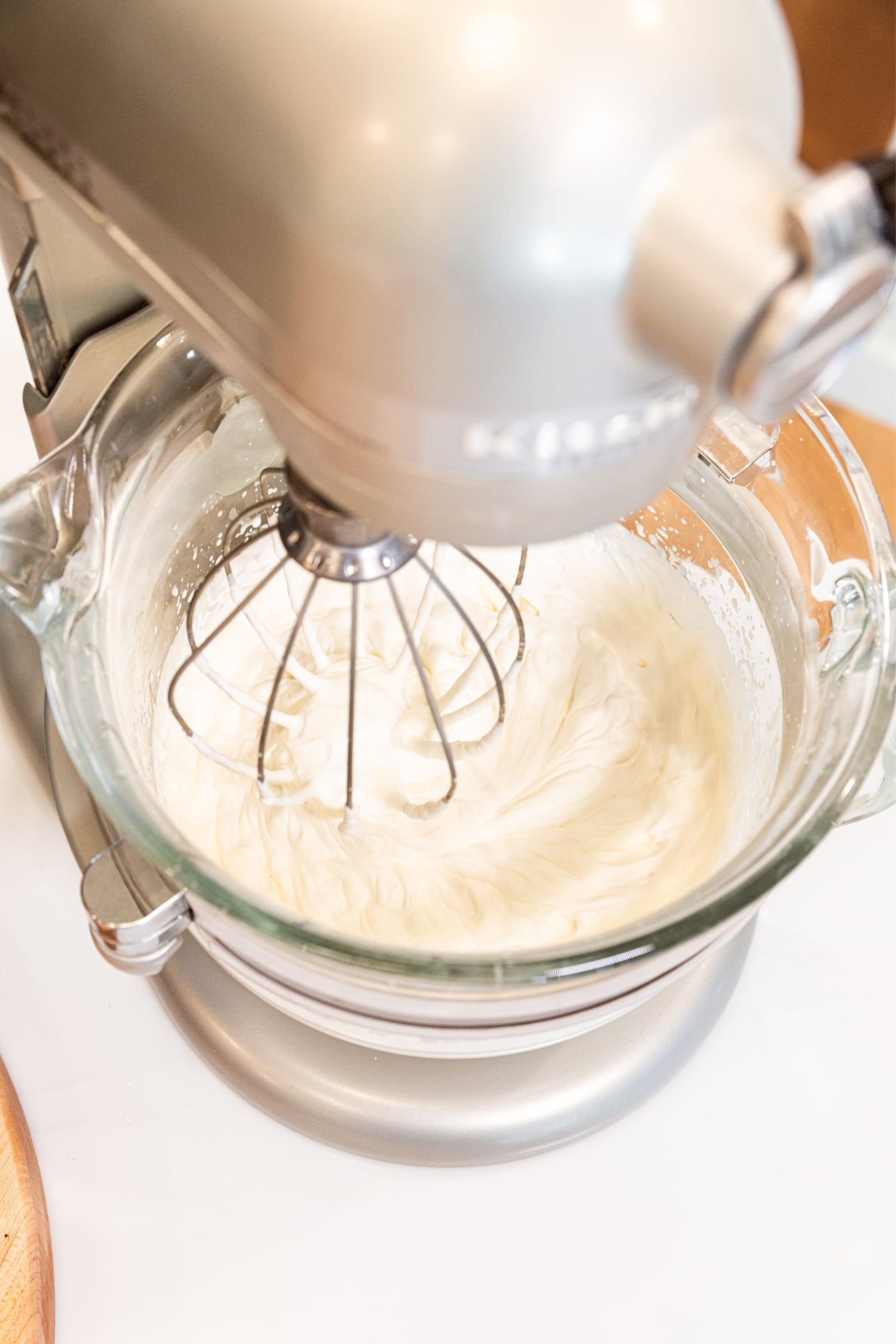 Glass bowl attached to stand mixer with whisk attachment mixing together heavy cream and powdered sugar into whipped topping for cake.