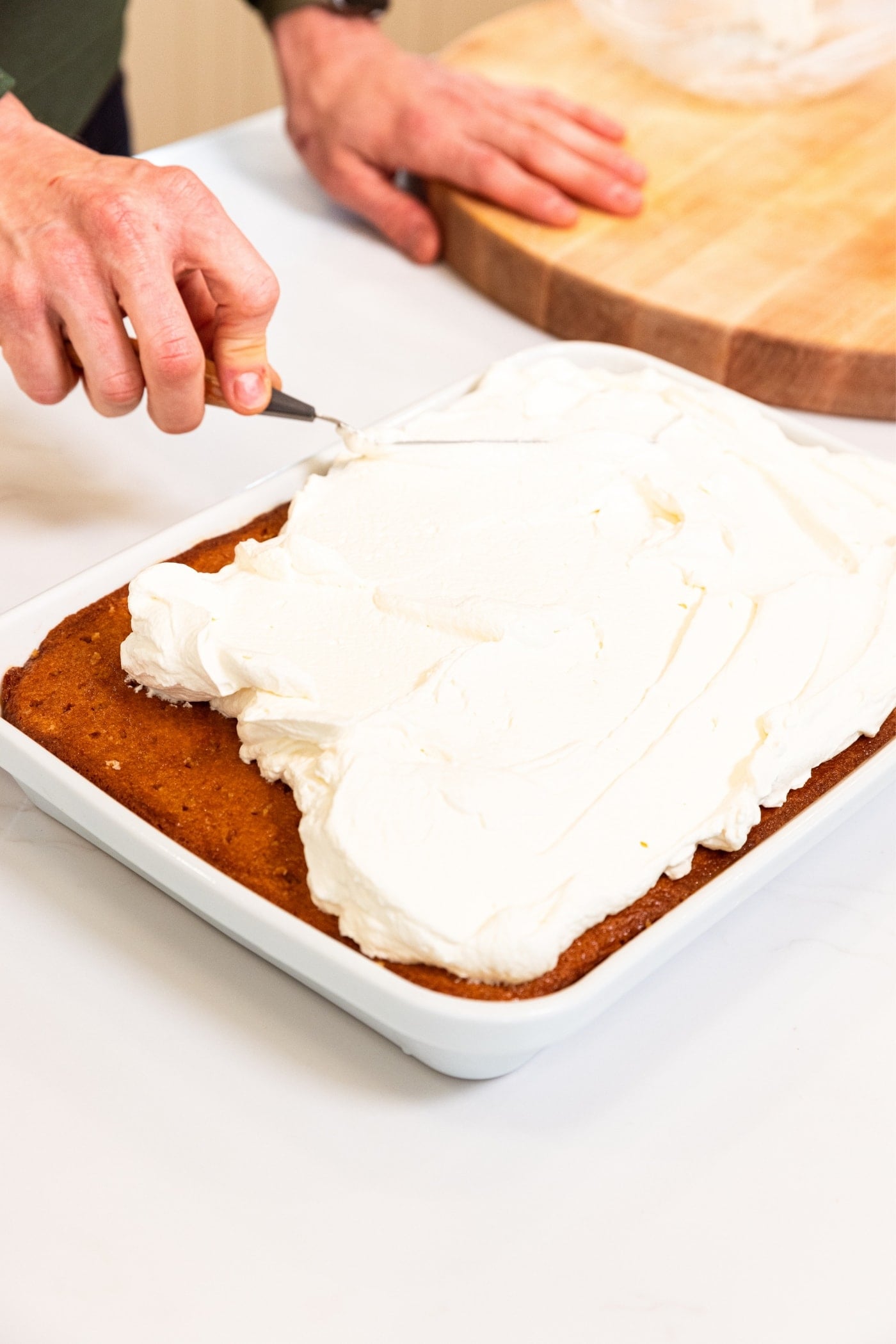 White baking dish filled with tres leches cake with hand spreading whipped topping using offset spatula.