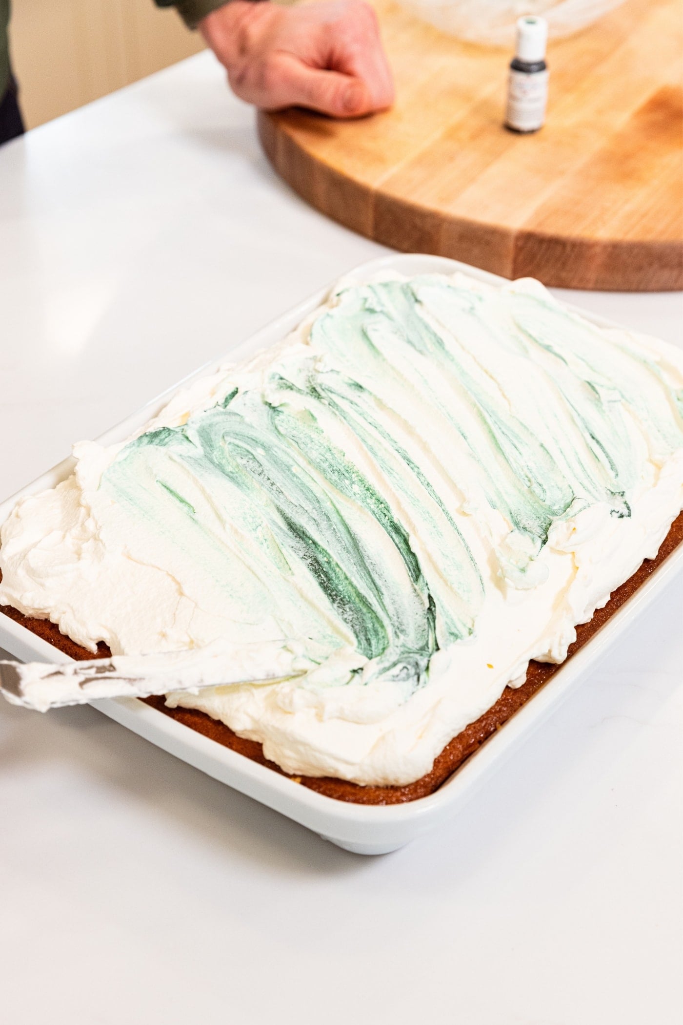 Hand spreading whipped topping on top of tres leches cake in white baking dish with green swirls in frosting.