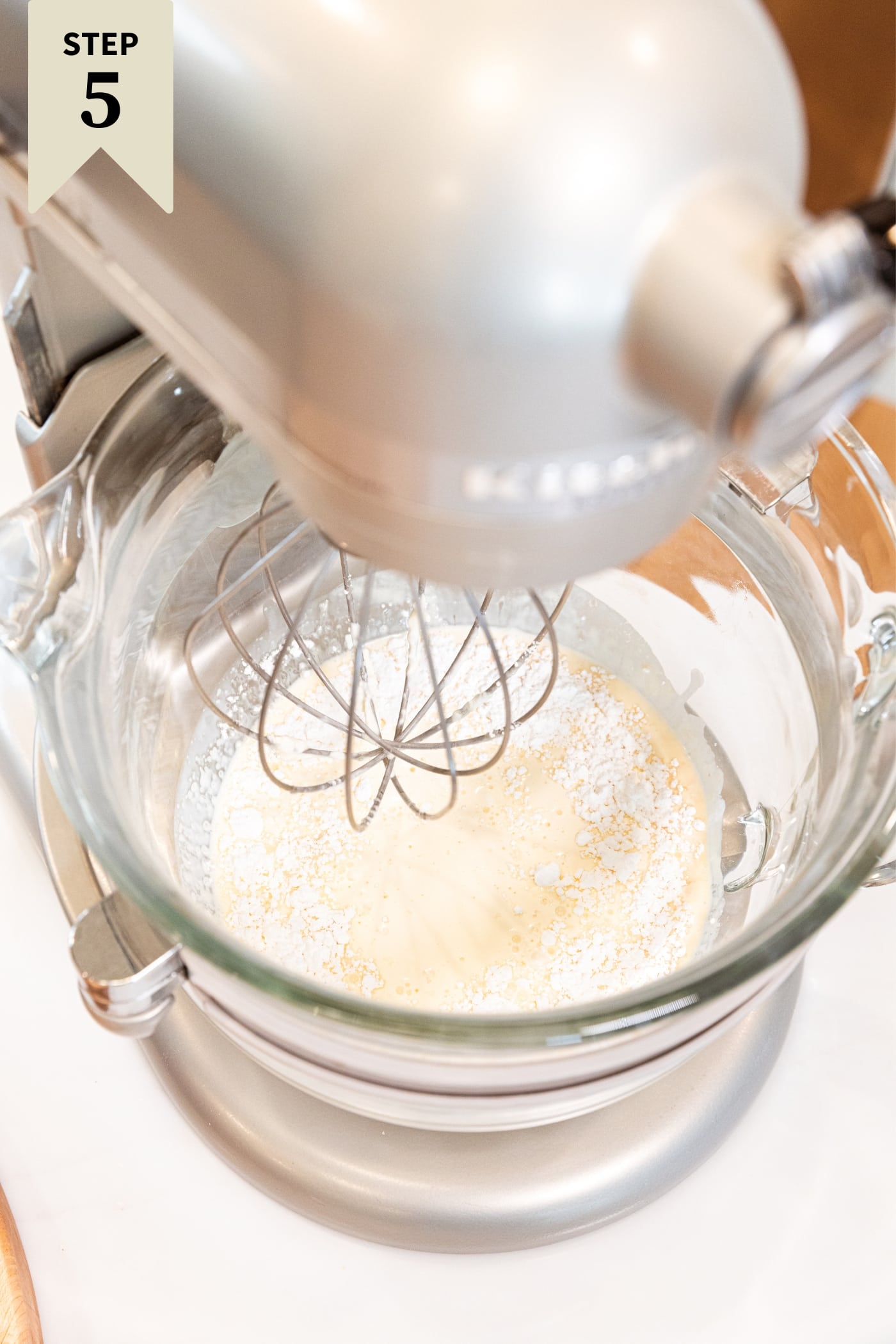 Large glass mixing bowl attached to stand mixer mixing together heavy cream with powdered sugar with whisk attachment.