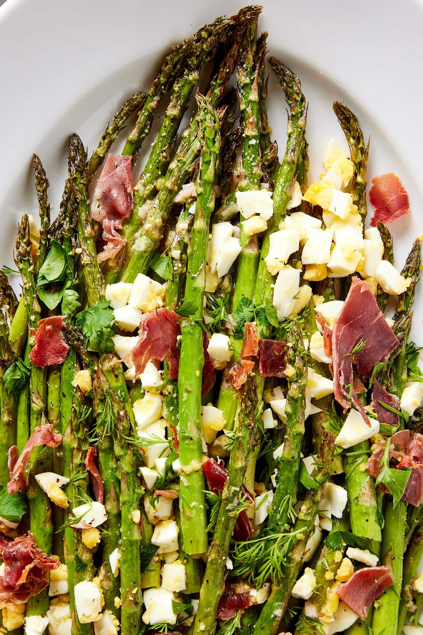 White platter filled with pieces of roasted asparagus with chopped hardboiled eggs and prosciutto on top with a sprinkling of herbs.