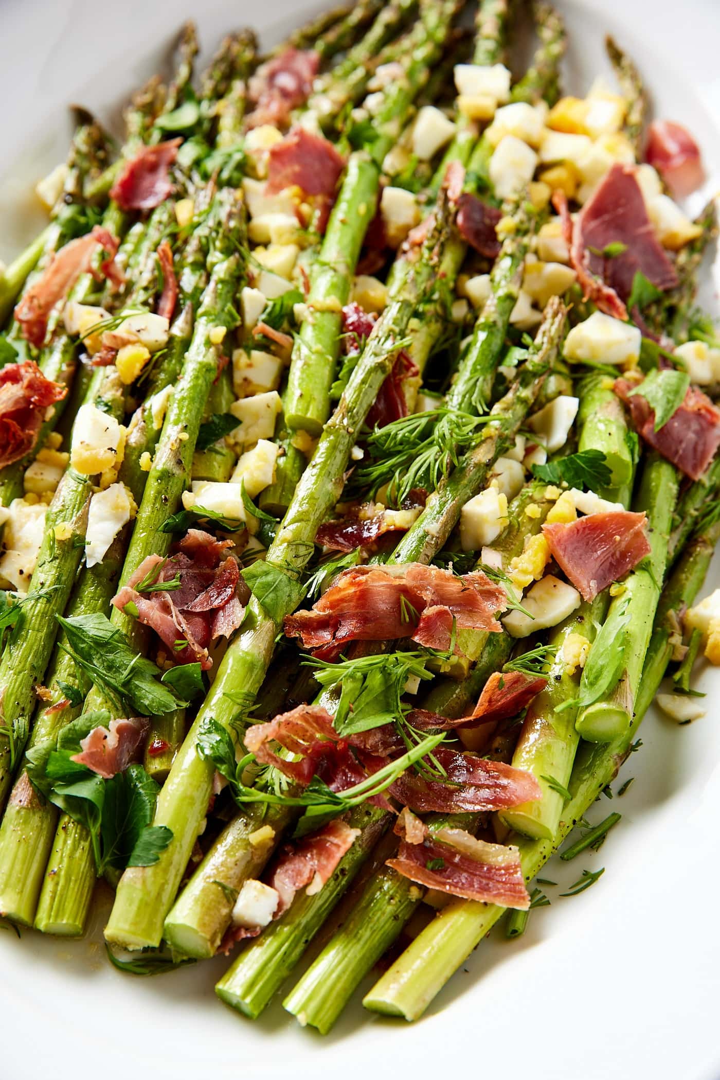 White bowl filled with pieces of roasted asparagus with chopped prosciutto and hardboiled eggs with herbs all over the top.