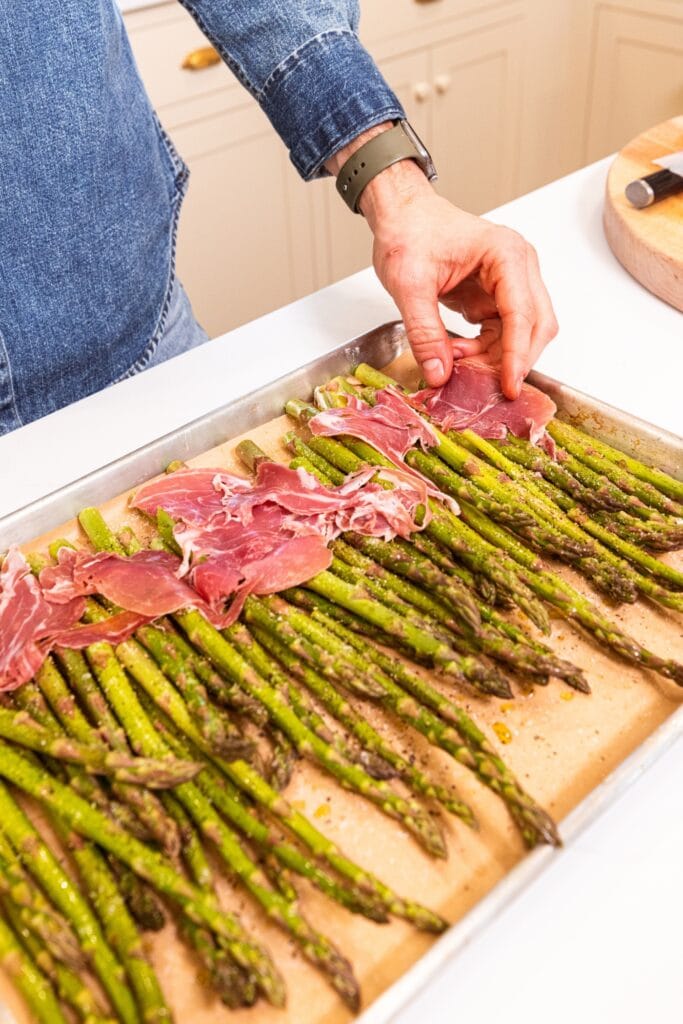 Hands laying pieces of prosciutto on top of pieces of asparagus on a parchment-lined baking sheet.