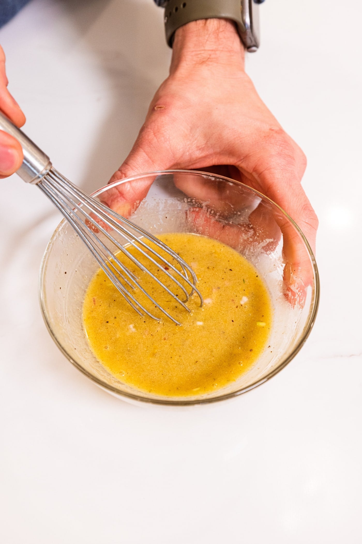 Hand stirring together vinaigrette in a small bowl on white surface.