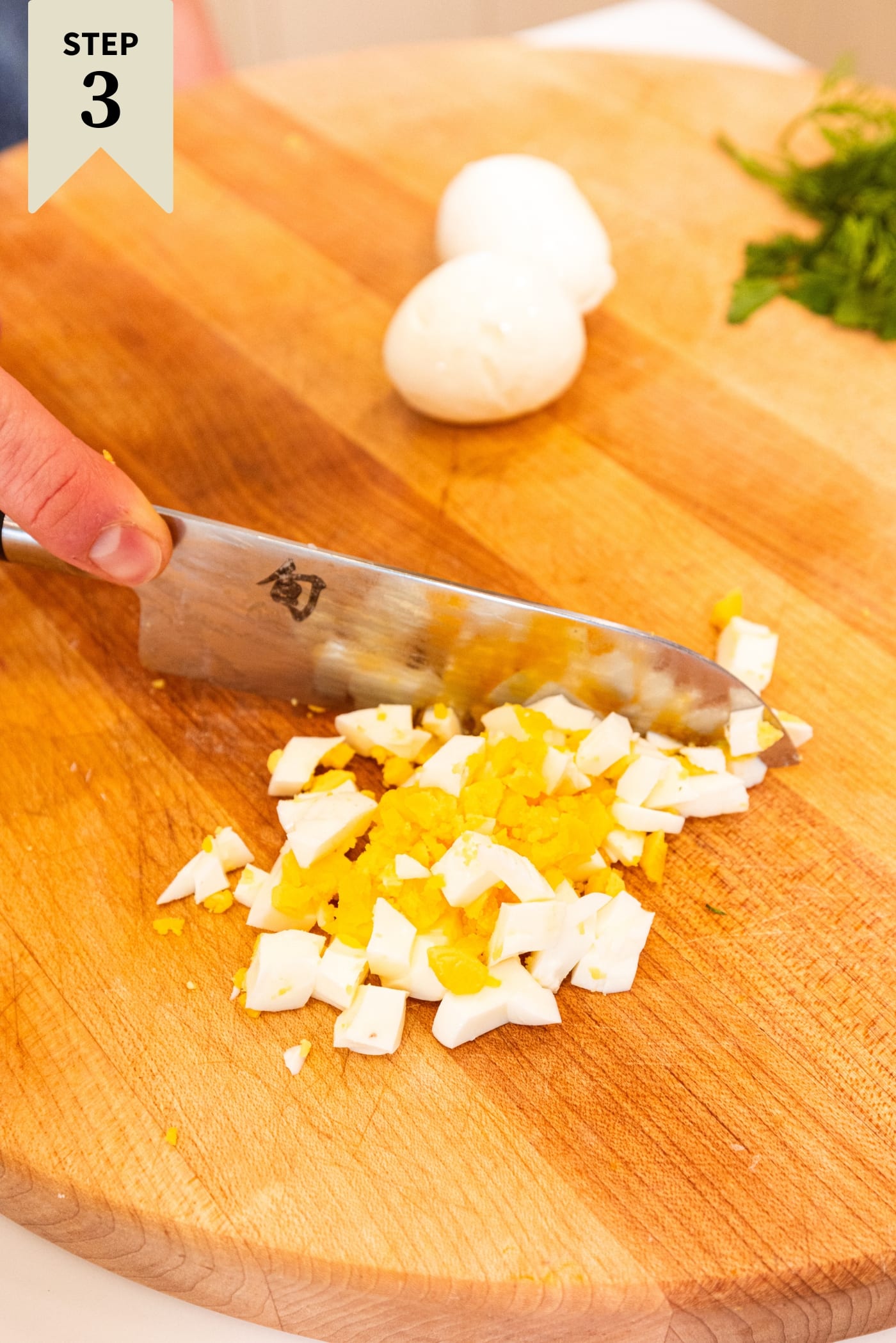 Hand using knife to slice hardboiled eggs into small pieces on wooden cutting board.