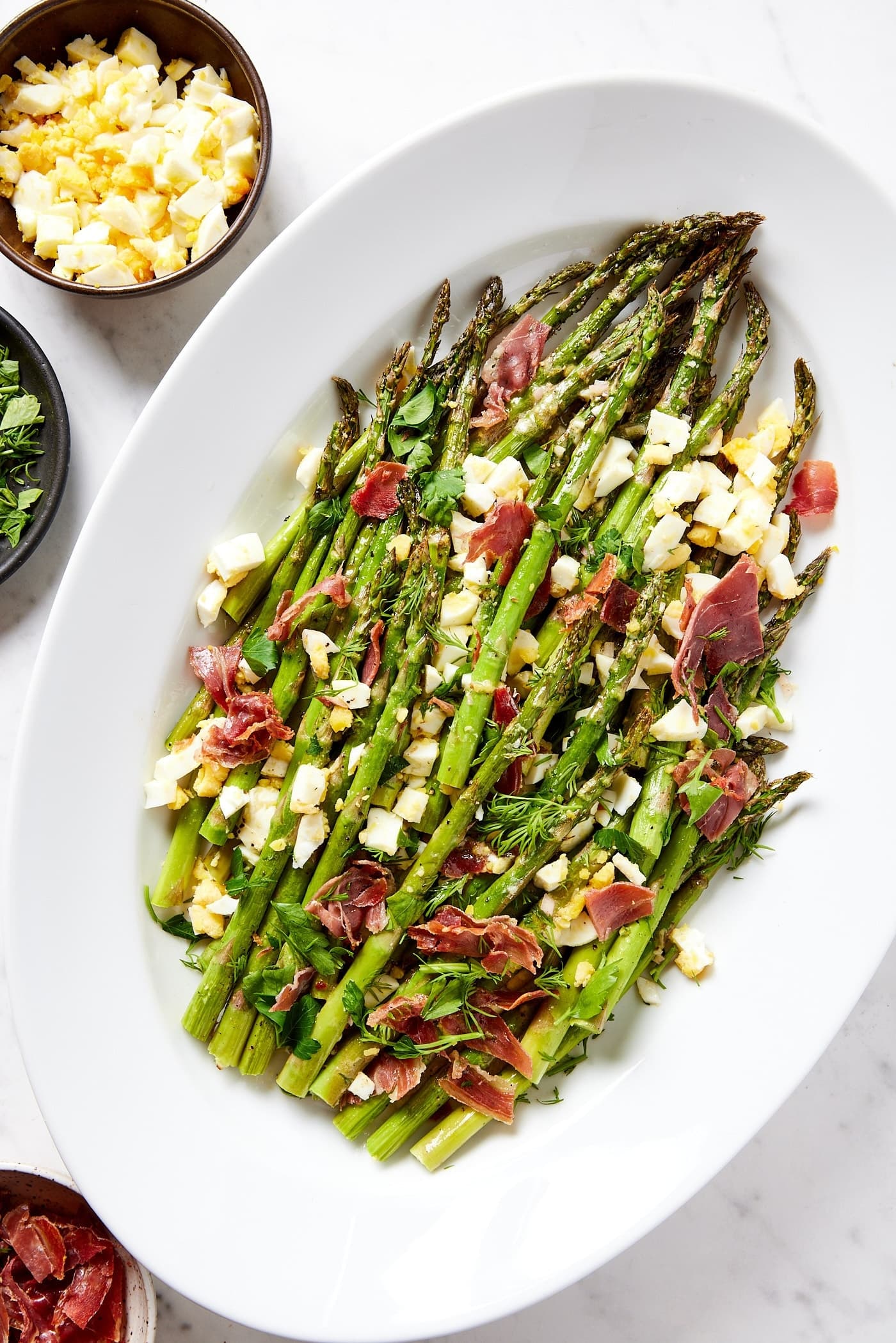 White platter filled with roasted pieces of asparagus topped with prosciutto and hardboiled eggs on white marble surface.