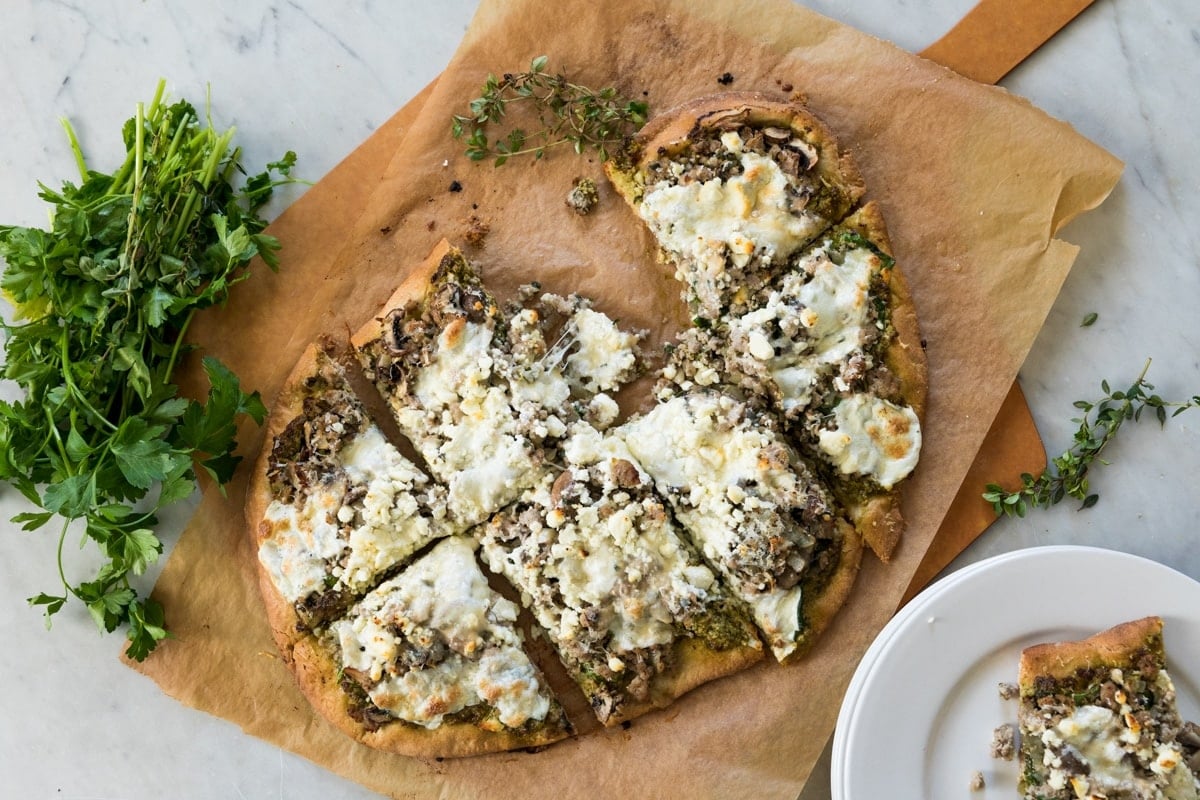Top down view of sliced pizza sitting on wood board with piece of parchment underneath with one slice sitting on a white plate all after coming out of the oven from baking