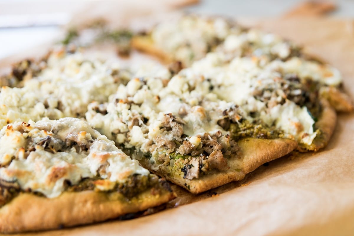 Slice of spinach pizza sitting on brown piece of parchment paper topped with sausage and crumbly feta cheese after coming out of the oven