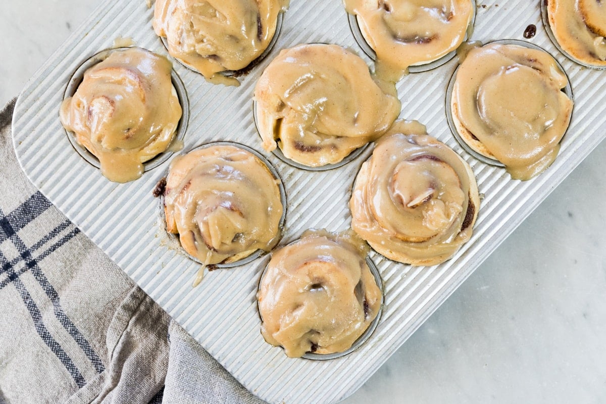 Top down view of silver muffin in containing frosted cinnamon roll muffins with towel on top of marble surface