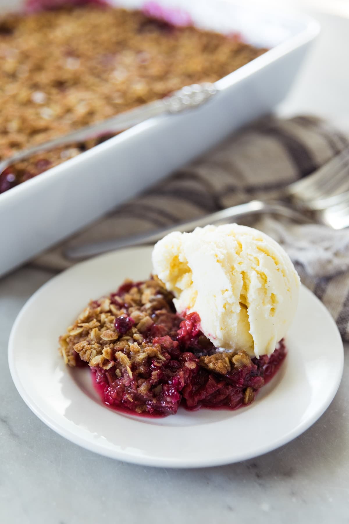 White plate filled with serving of rhubarb and berry crisp with oats on top with large scoop of vanilla ice cream on top with baking dish in background