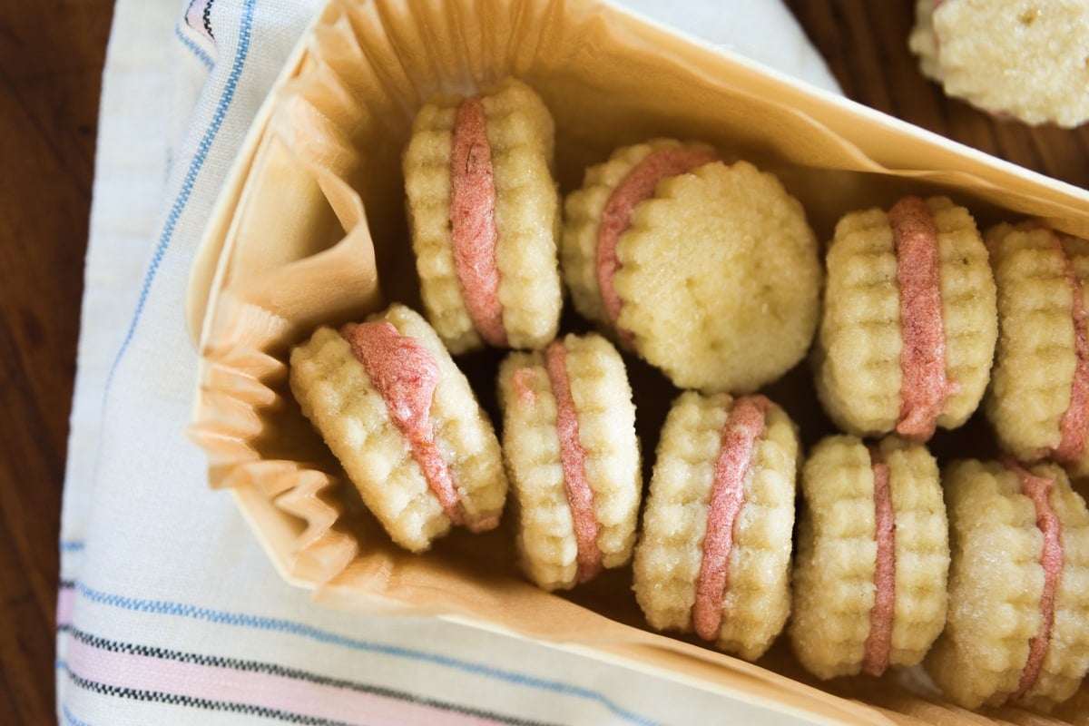 Top down view of rhubarb wafer cookies with light pink filling sitting in a parchment lined treat box one top of white dish towel