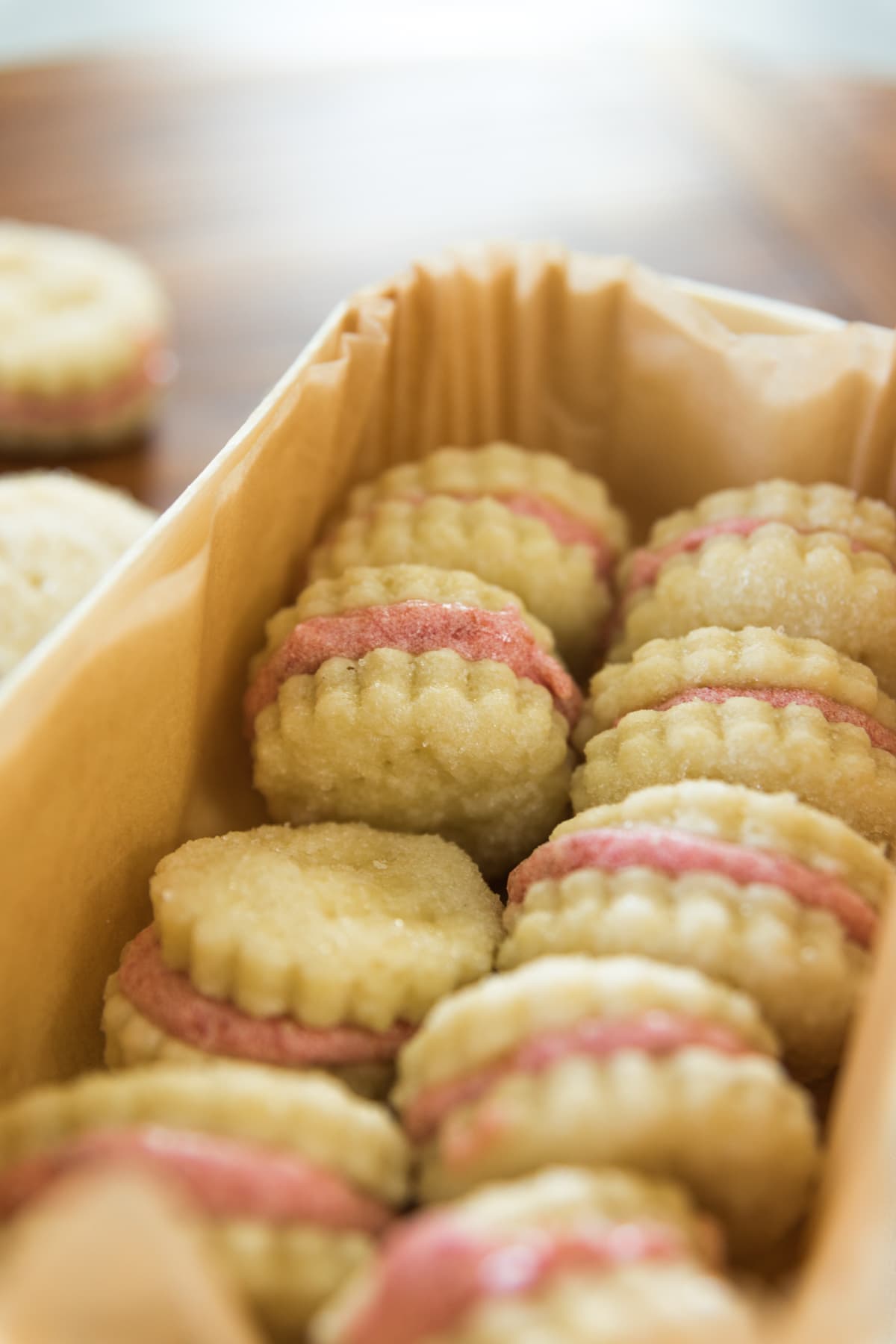 Light brown sandwich cookies with light pink frosting filling sitting in box lined with parchment paper one wood surface