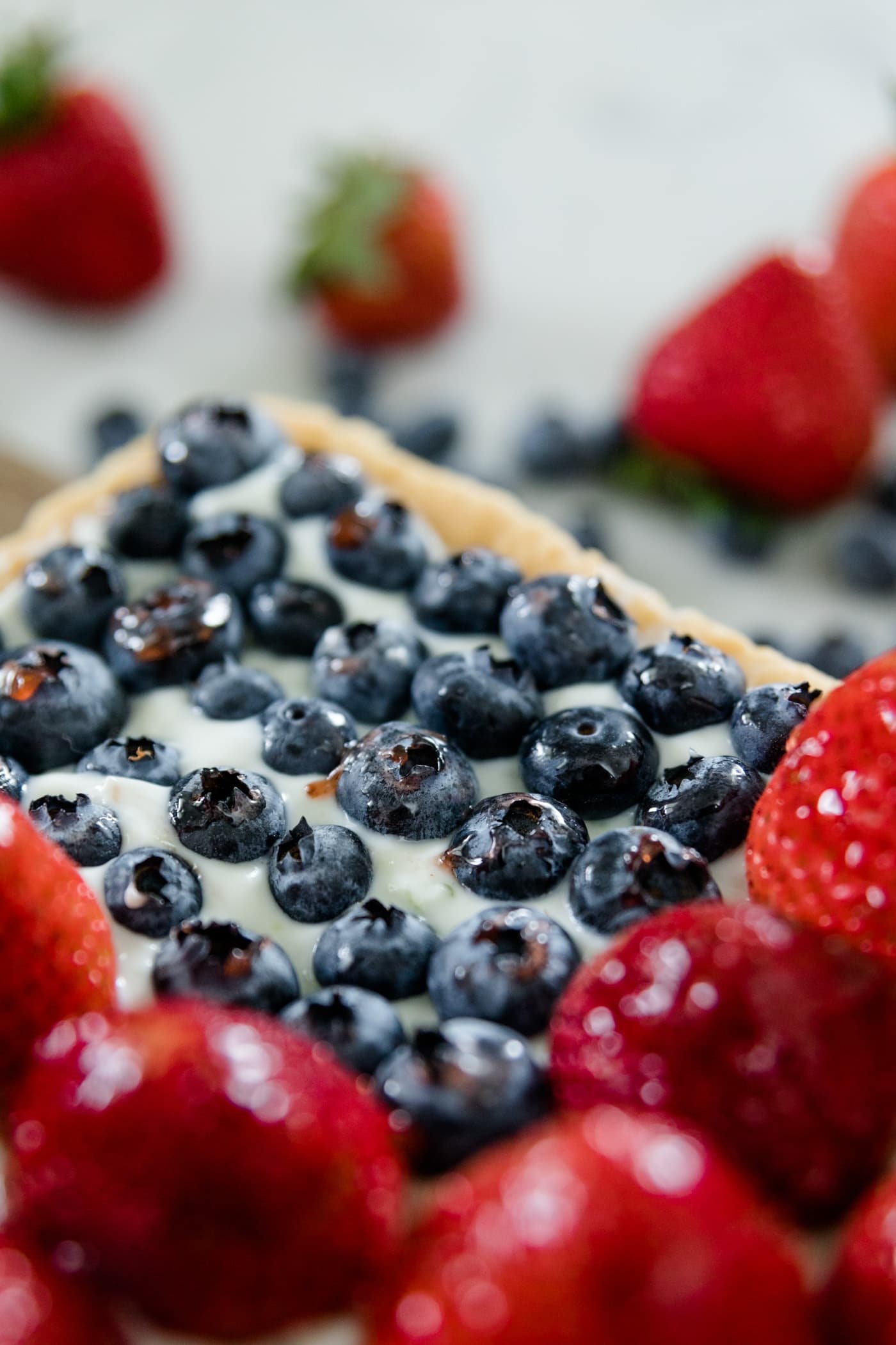 Strawberries and blueberries on a cookie tart with yogurt filling with extra strawberries and blueberries in background on a marble surface
