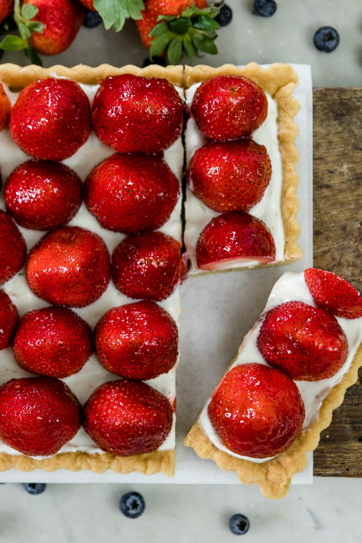 Top down view of a piece of strawberries on top of a yogurt filling and a cookie tart on a wood cutting board on a marble surface
