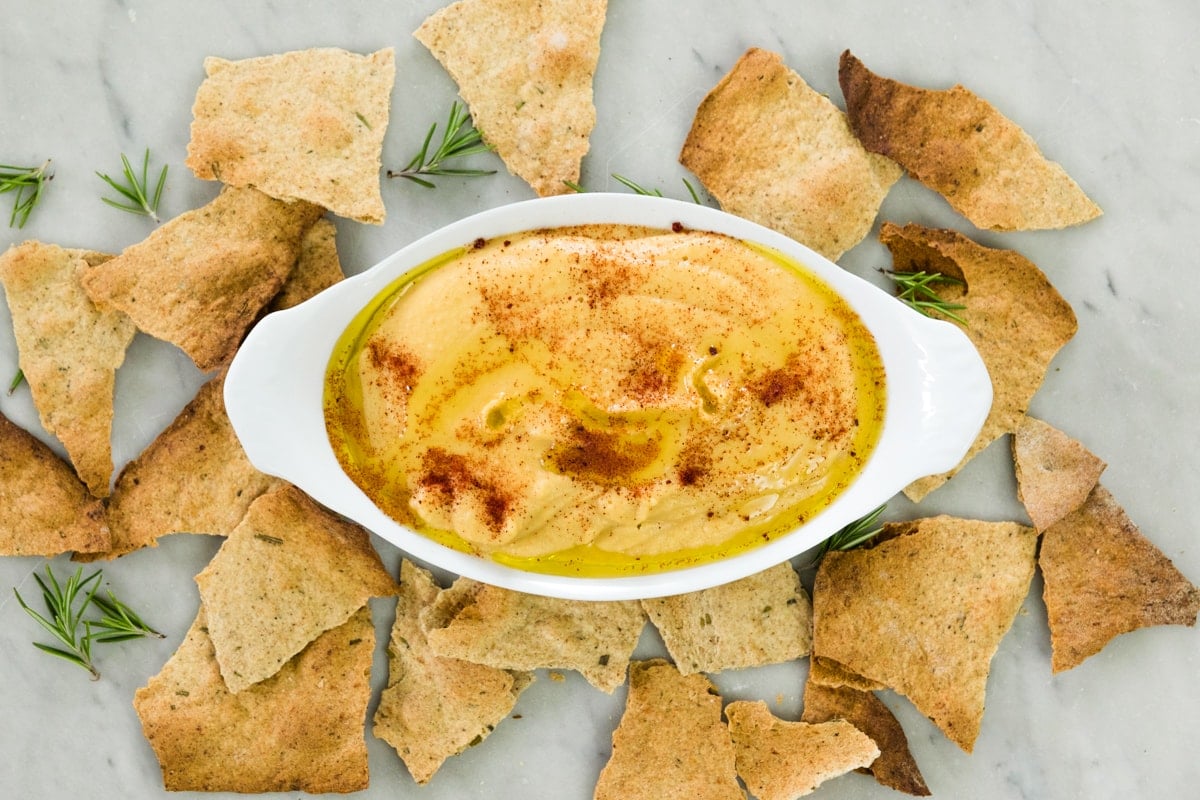 Top down view of white oval bowl filled with light brown colored traditional hummus topped with paprika and oil sitting on marble surface with broken crackers all around
