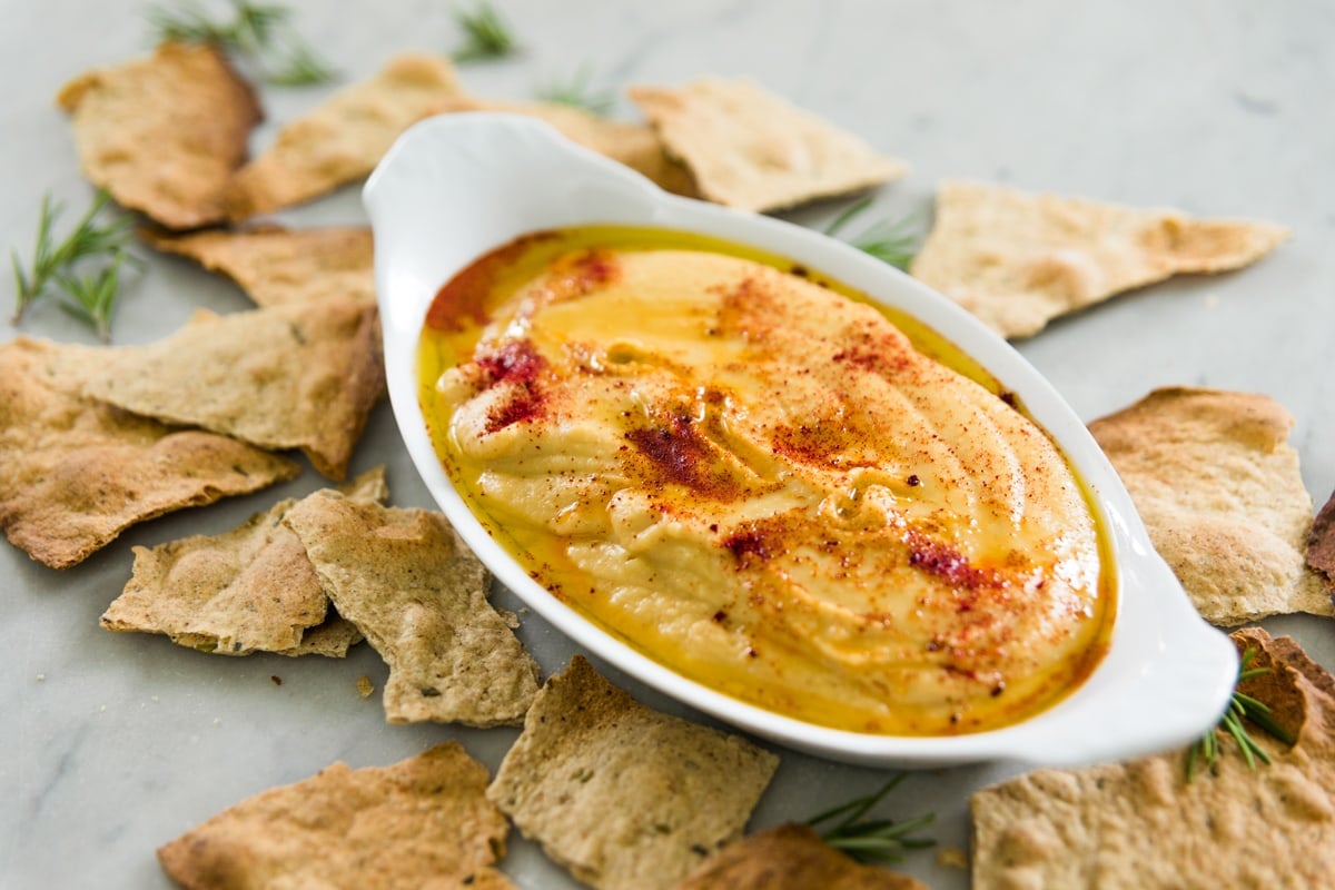 White oval bowl filled with traditional hummus topped with oil and paprika with rustic herb crackers all around on marble surface