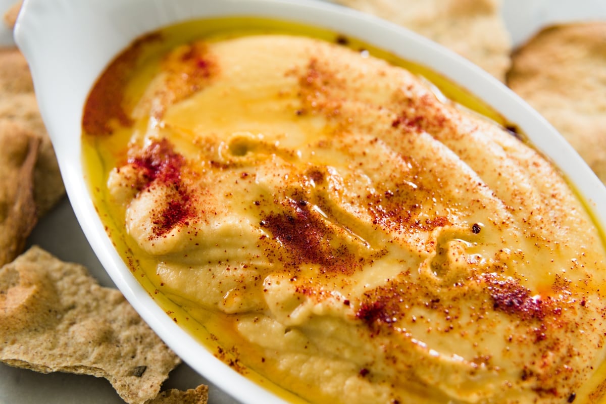 Close up view of white oval serving bowl filled with traditional hummus with paprika on top with rustic crackers all around