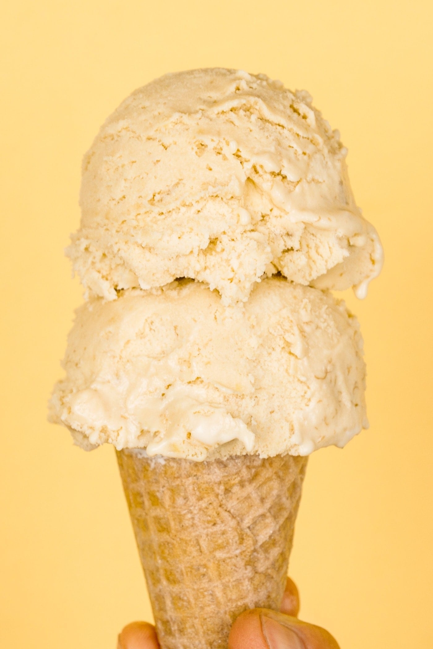 Two scoops of banana cream pie ice cream in ice cream cone held by a hand with light peach colored background