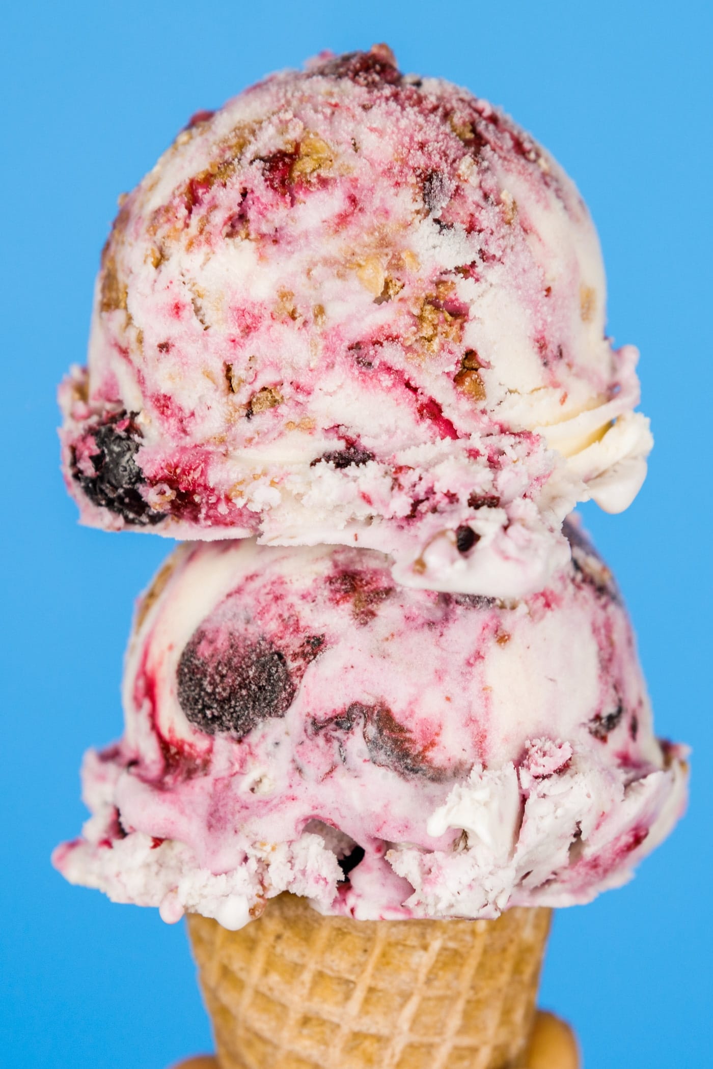 Two scoops of blueberry crumble pie ice cream with swirls of blueberry and homemade streusel on top of a cone with a bright blue background