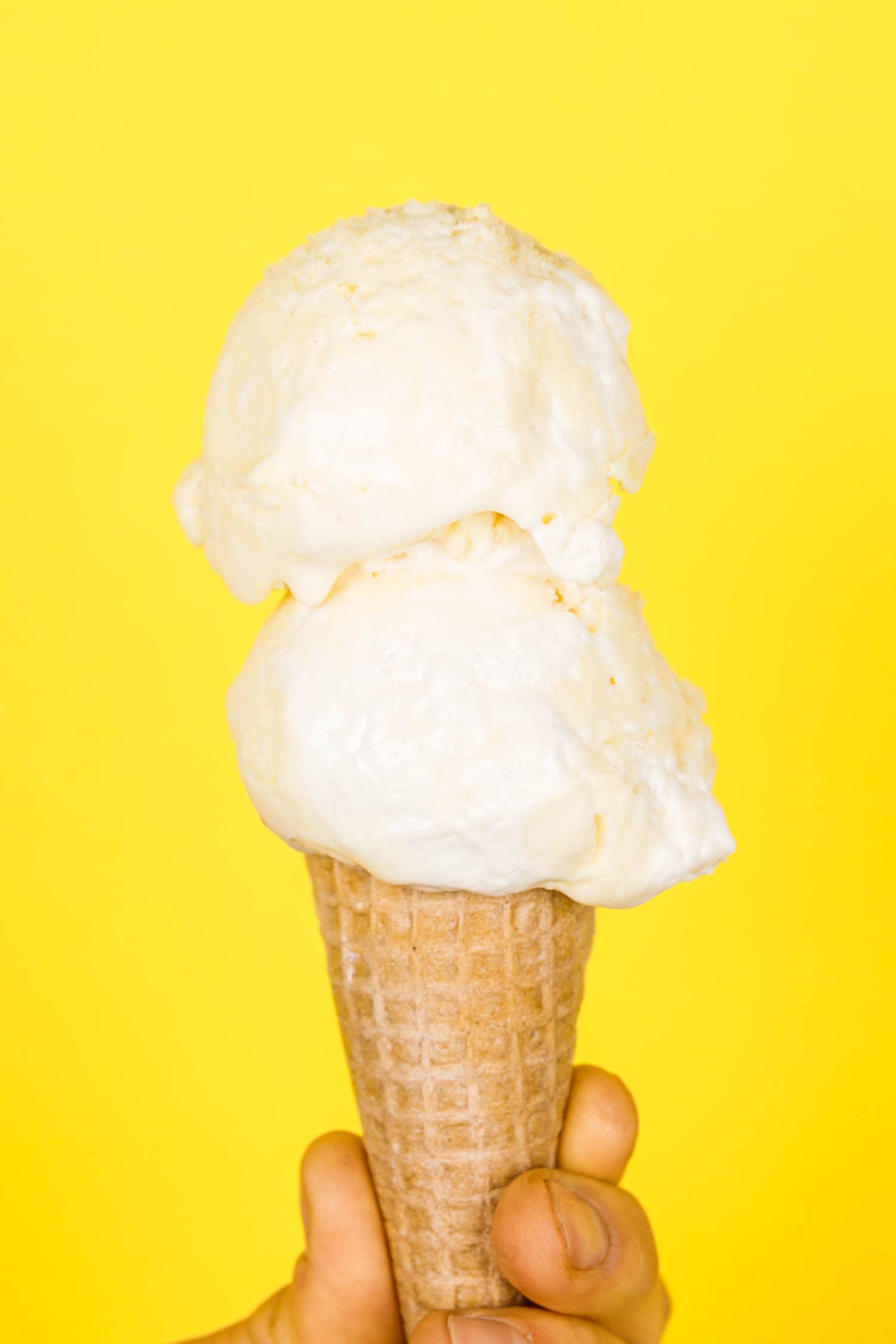 Two scoops of lemon meringue pie ice cream on a cone being held in front of a bright yellow background