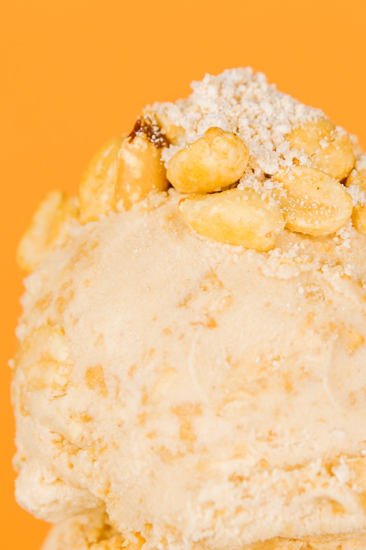 Two scoops of peanut butter pie ice cream on an ice cream cone with peanuts and fillings on top in front of orange background