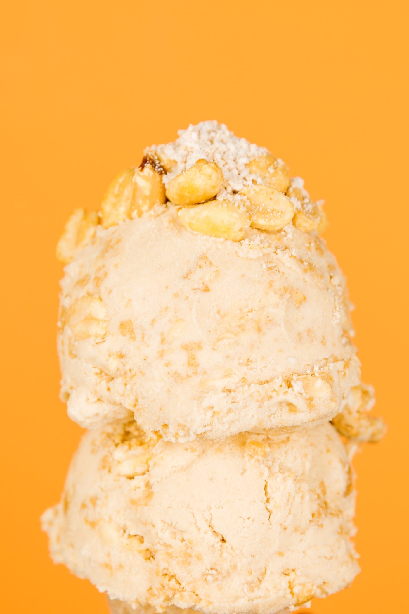Two scoops of peanut butter pie ice cream on an ice cream cone with peanuts and fillings on top in front of orange background