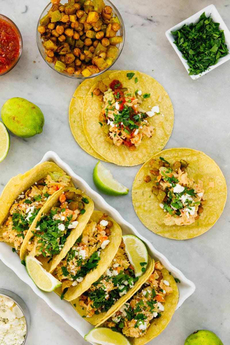 Top down view of chickpea tacos sitting on marble surface with various toppings scattered all around including limes, salsa, and cheese