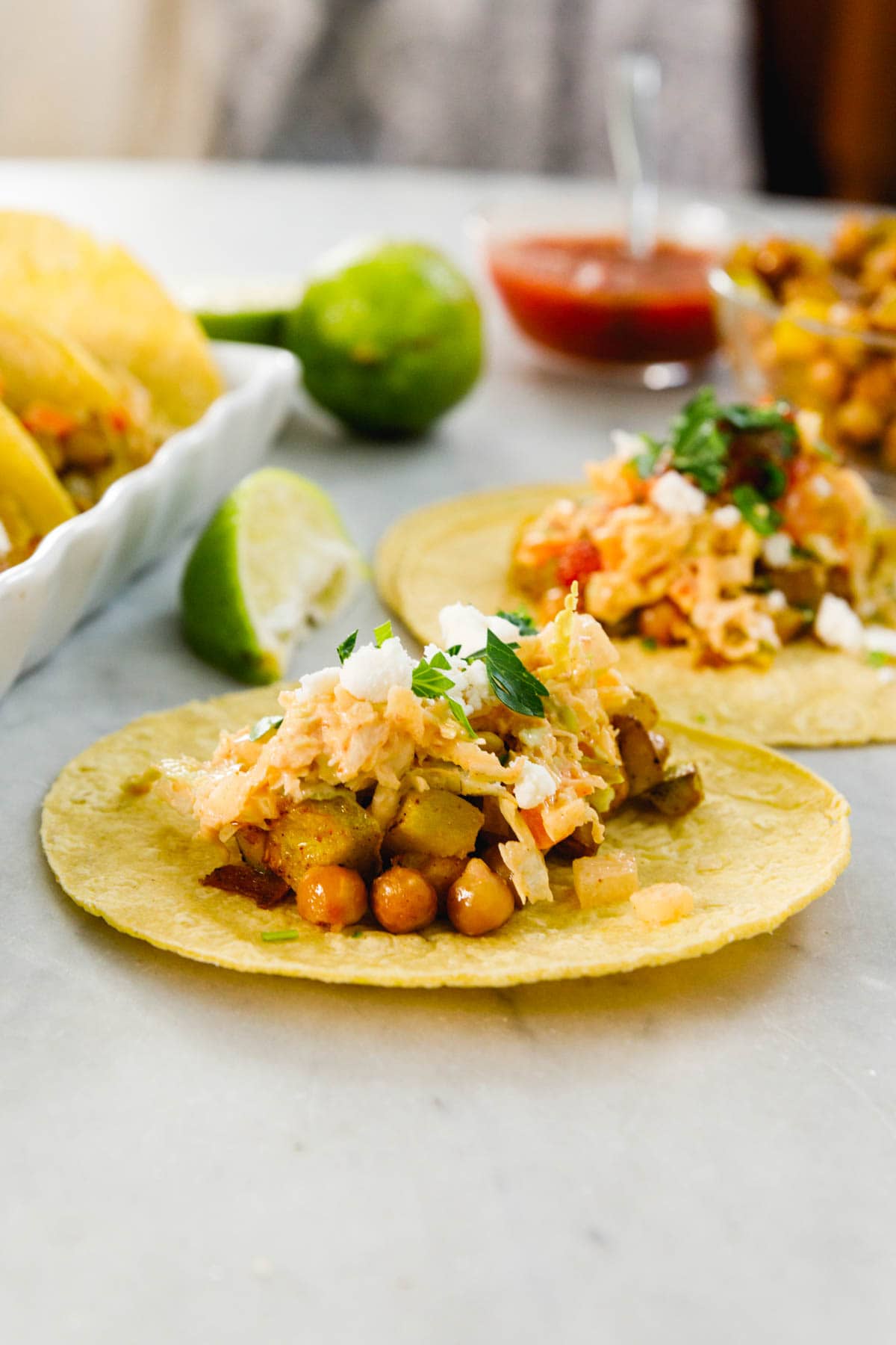 Corn tortilla sitting on marble surface with chickpea tacos mixture on top along with squash and cilantro with more tacos and toppings in background