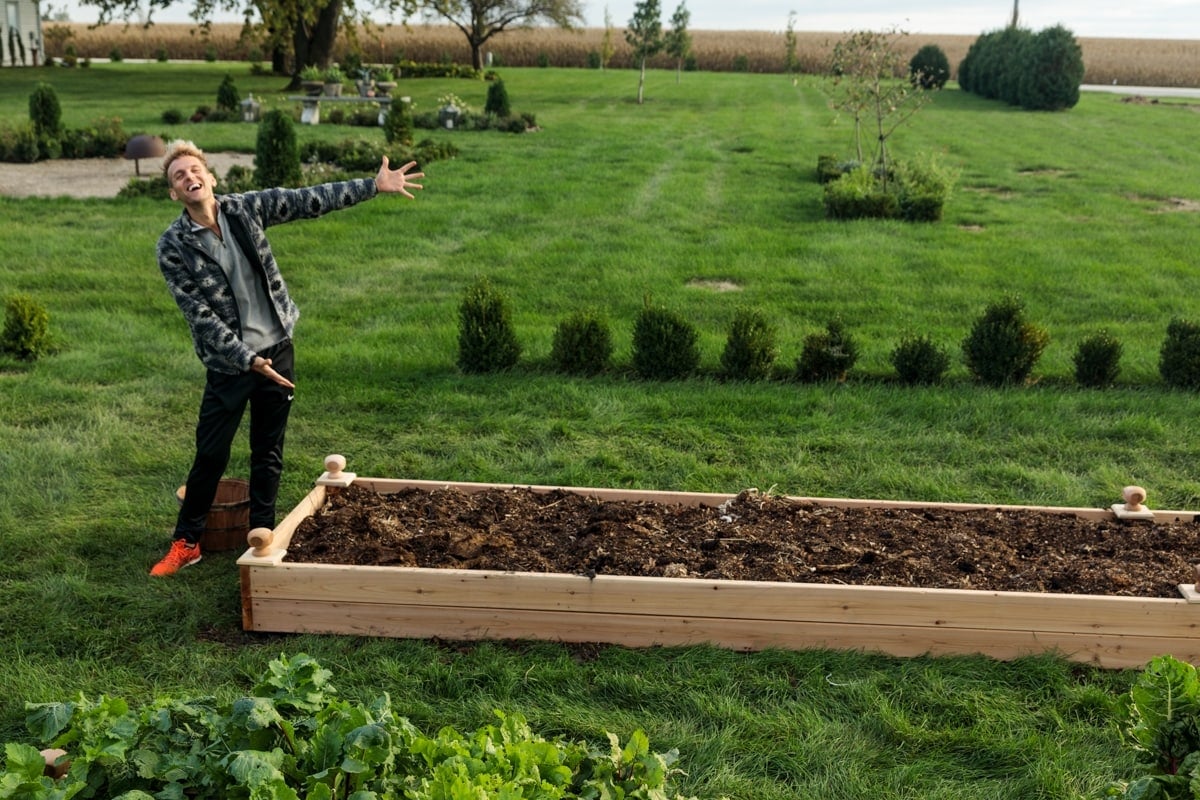 Man with blonde hair standing beside completed raised garden bed filled with dark brown soil with grass all around