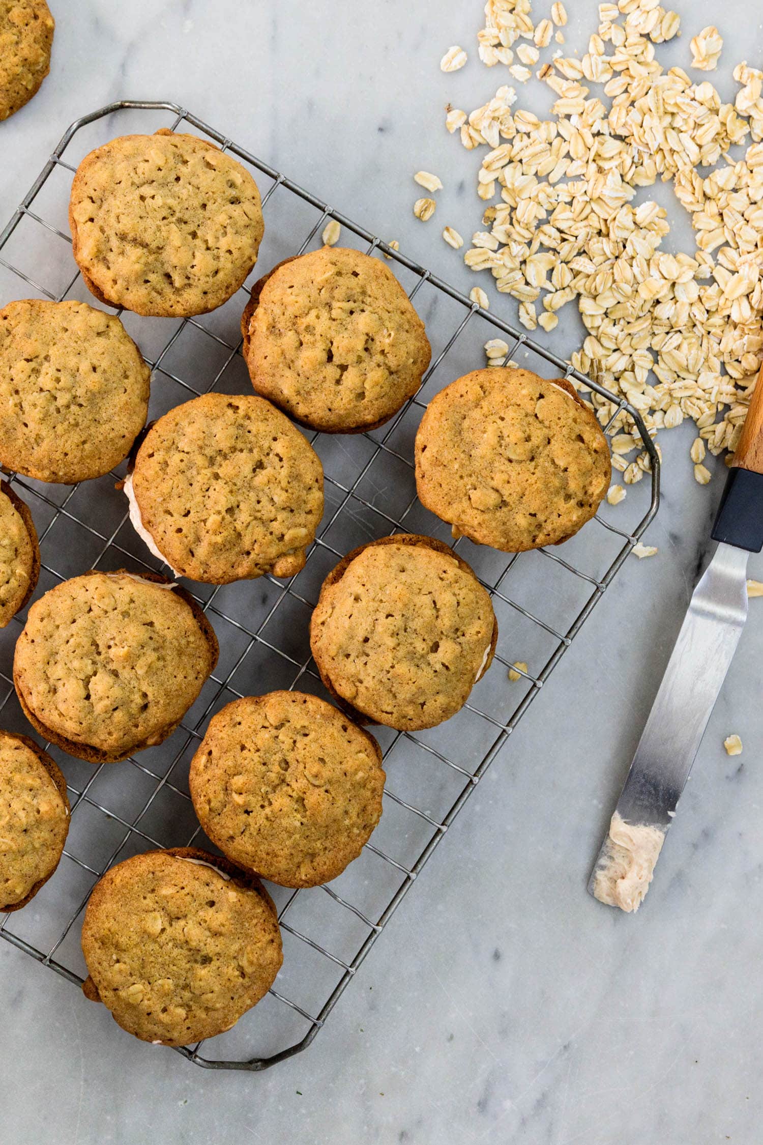Top down view of oatmeal spice creme-filled cookies all on a wire cooling rack on a marble surface with oats and knife with extra frosting