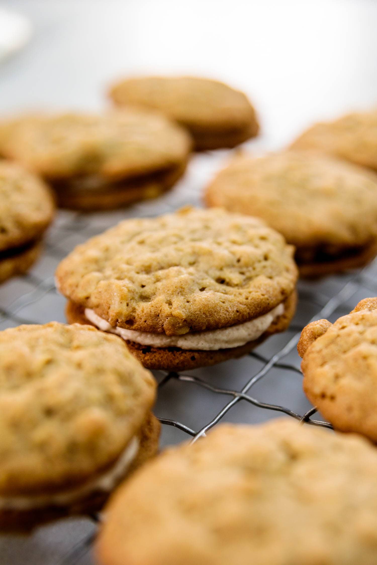 Two oatmeal spice cookies sandwiching a layer of white creme filling with multiple cookies in background all on a wire cooling rack on a marble surface