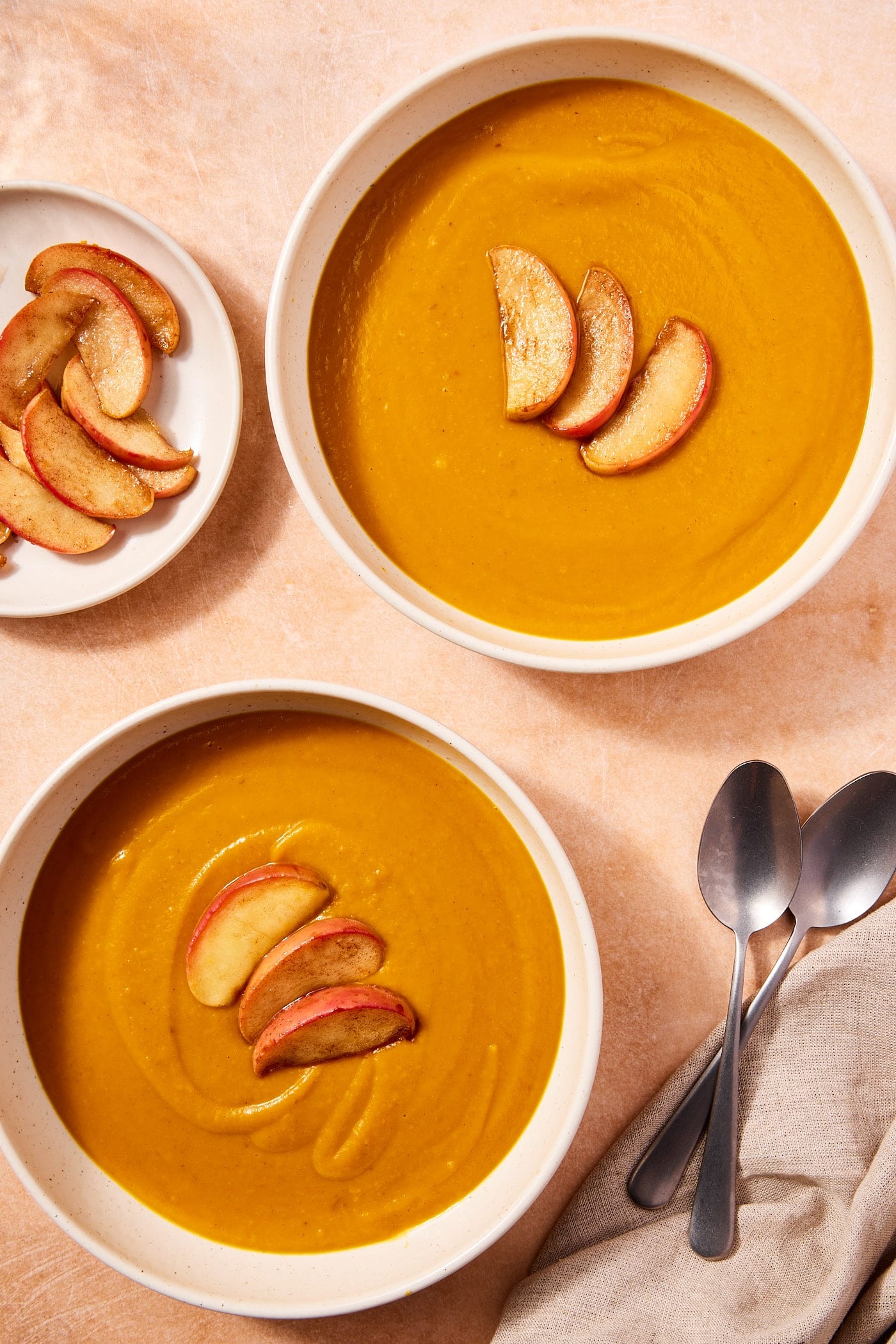 Two large bowls filled with squash soup with sliced and cooked apples to the side along with spoons.